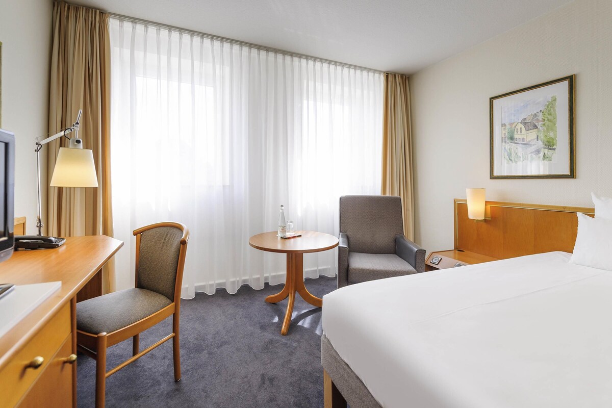 A hotel room with a bed, a desk with a lamp, two chairs, a small table, and a painting on the wall.