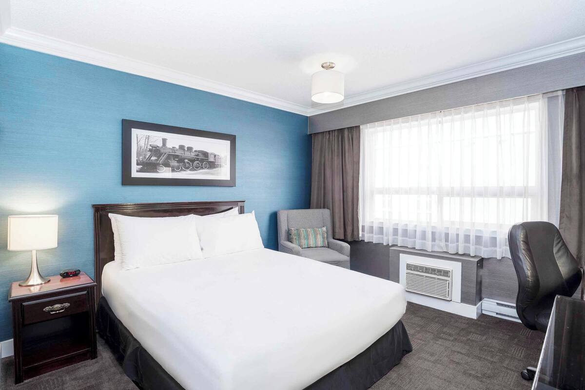 A hotel room with a large bed, a desk, a chair, a picture on the wall, and a window with curtains.