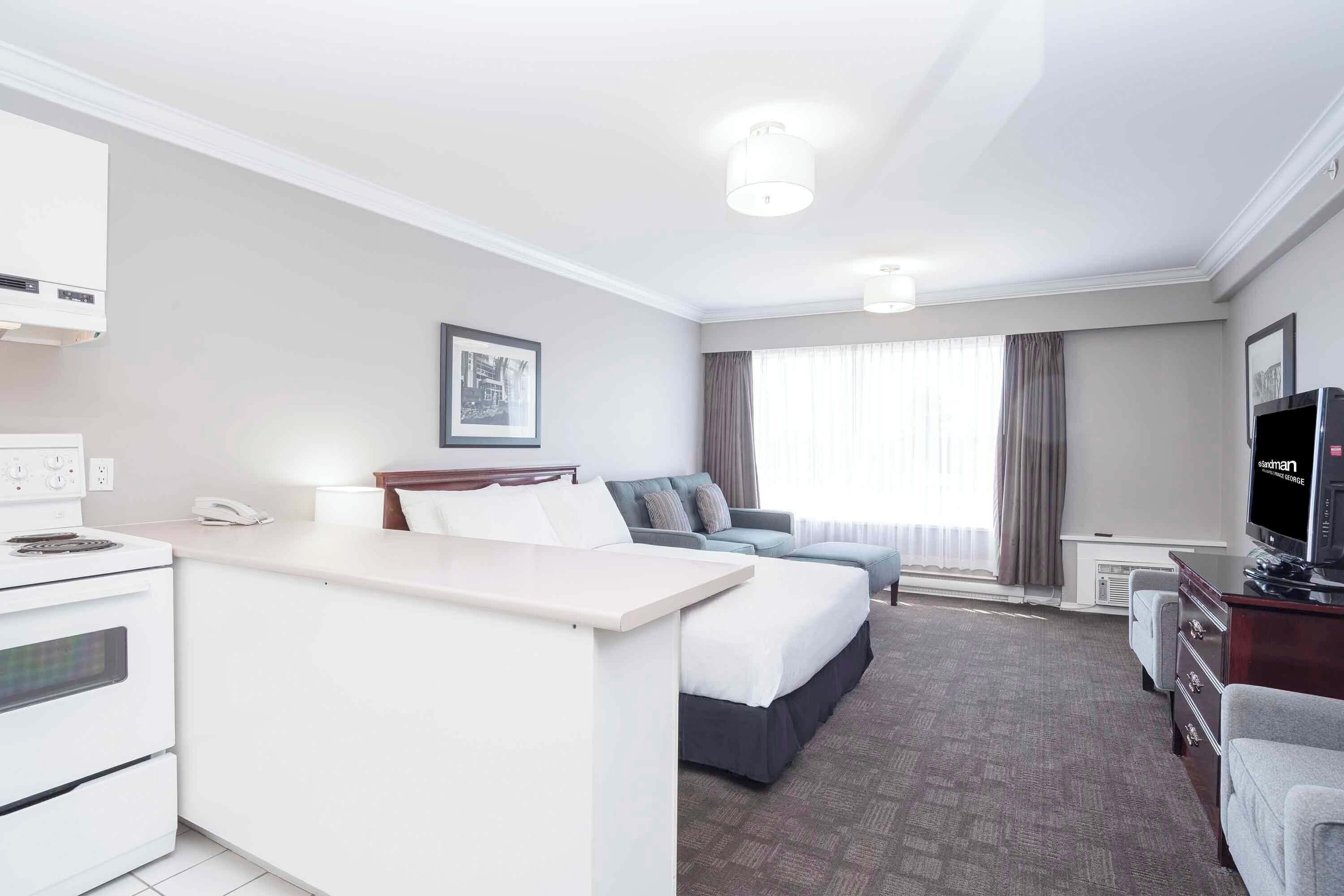 A hotel room with a kitchenette, a bed, a sofa, a TV, and a window with curtains.