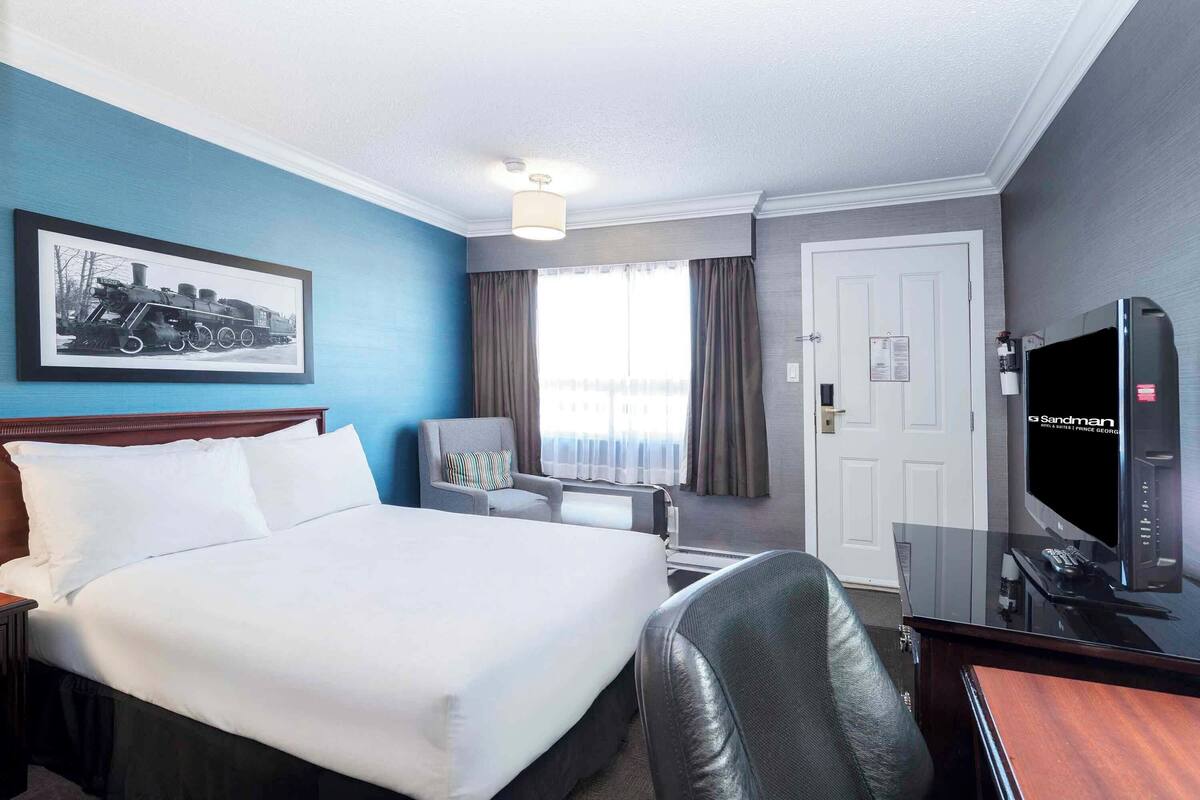 A hotel room with a large bed, a desk with a TV, a chair, and a window with curtains.