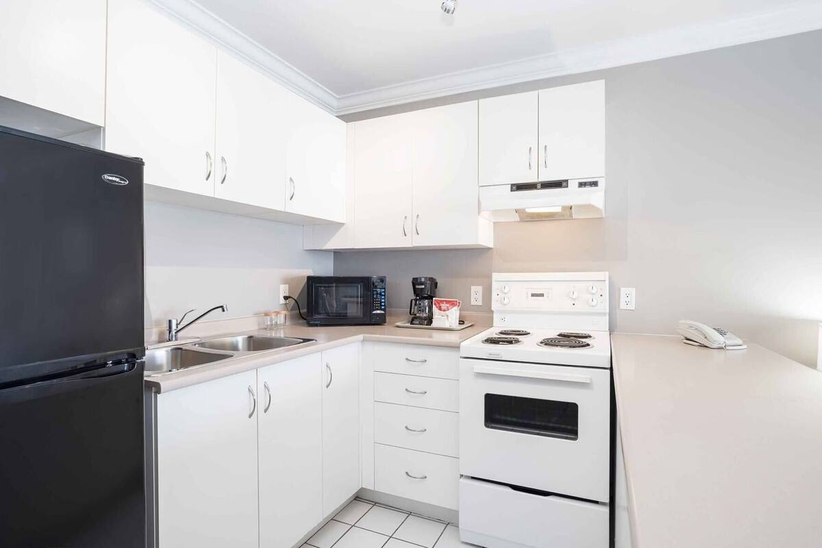 A compact kitchen with white cabinets, a black refrigerator, a microwave, a coffee maker, a stove, and a phone on the counter.