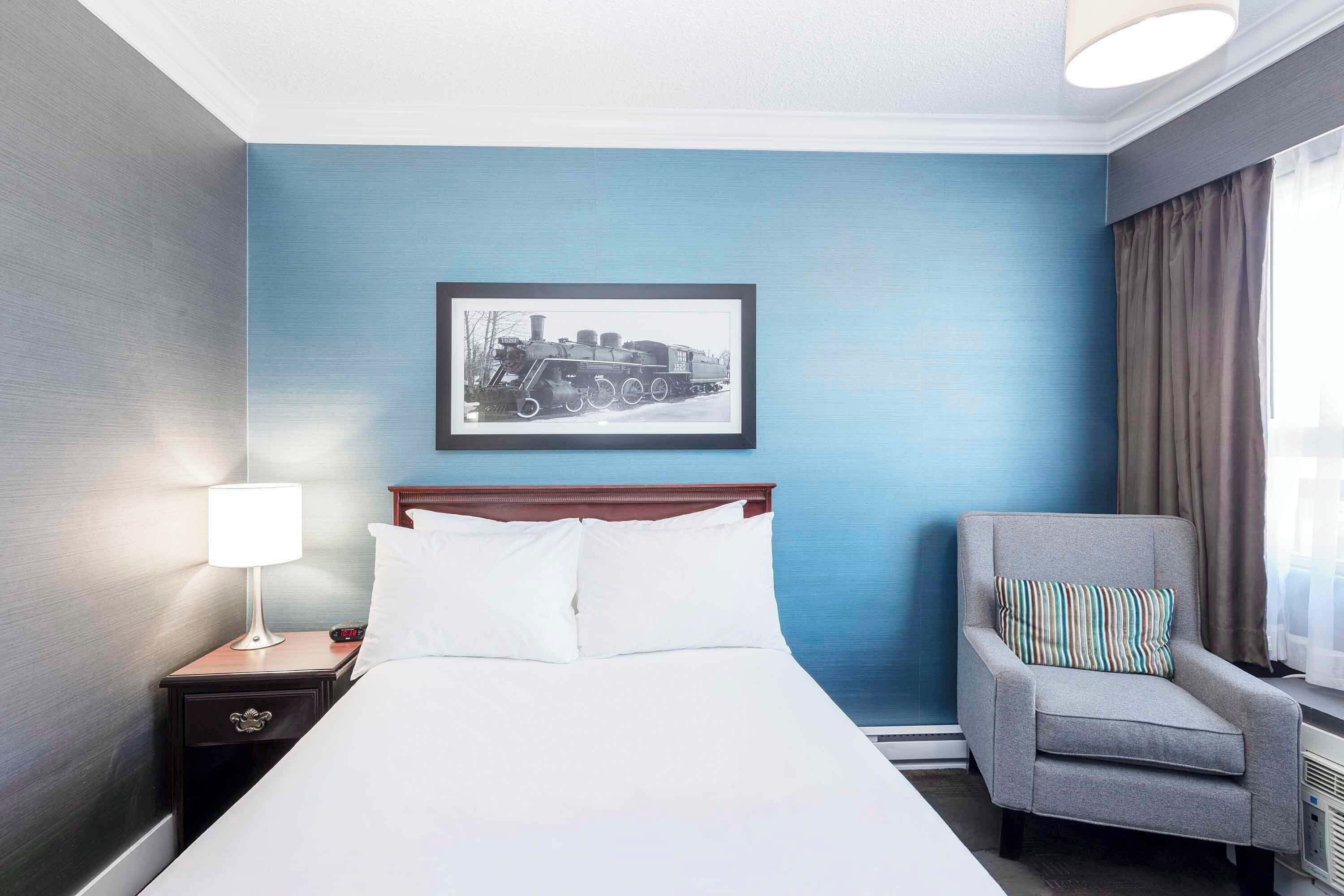 Une chambre d’hôtel avec un lit, une table de chevet avec une lampe, une chaise et un tableau encadré au mur.