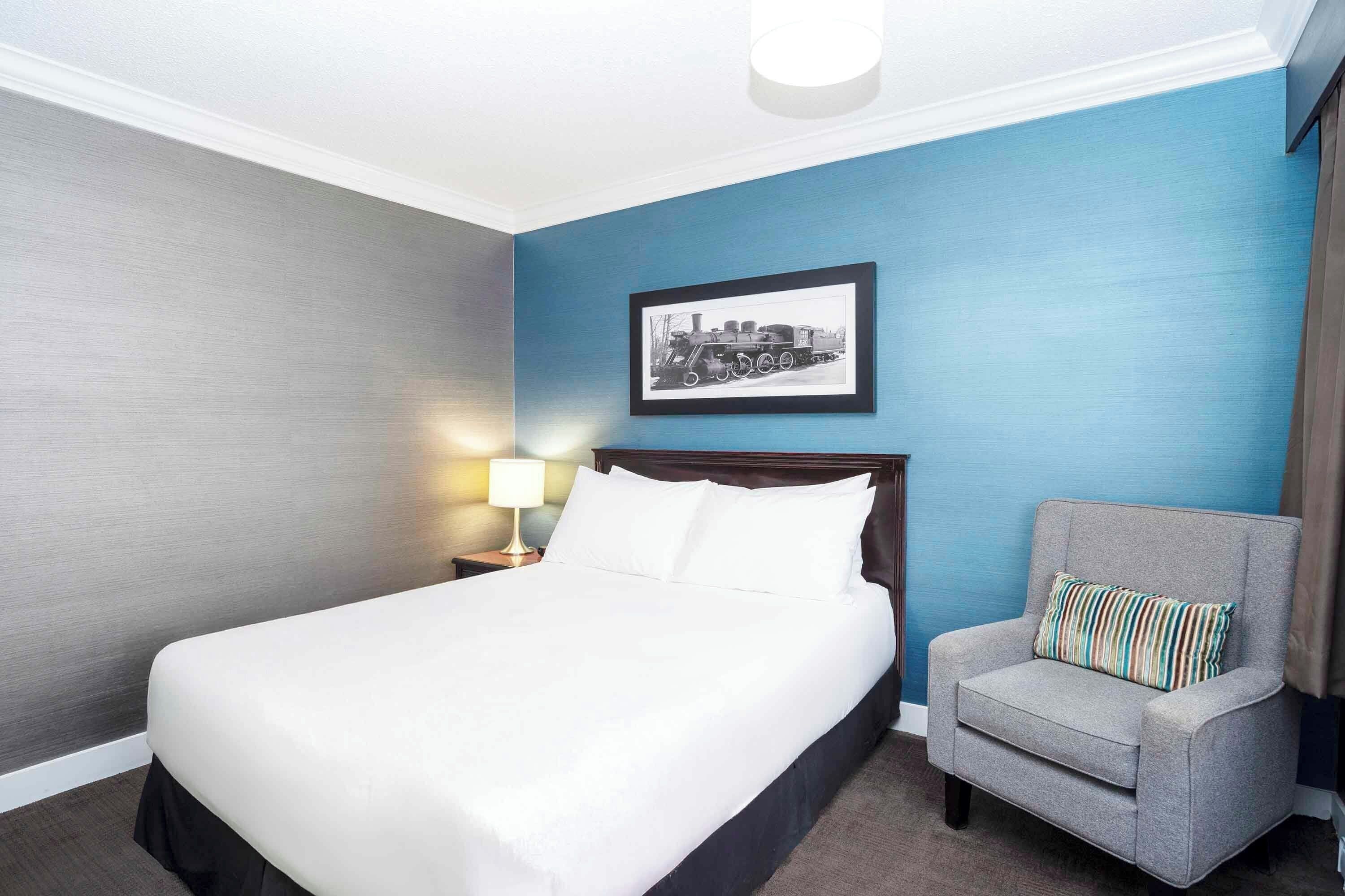A hotel room with a large bed, a gray armchair, a nightstand with a lamp, and a framed picture on the wall.