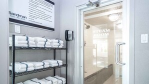 A hotel room entrance with a glass door leading to a steam room, a sign listing room amenities, and shelves with neatly folded towels. - Sandman Hotel & Suites Prince George (Prince George)