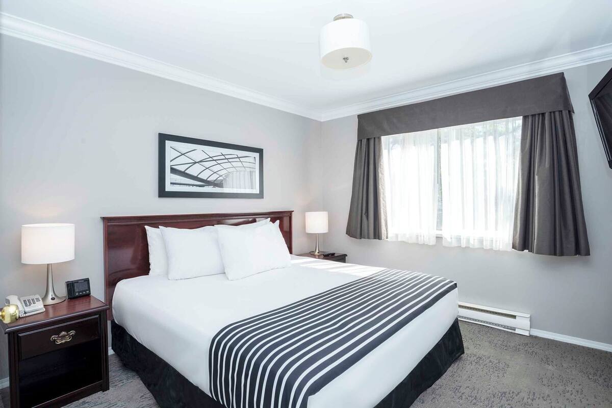 A hotel room with a large bed, two bedside tables, a nightstand, a framed picture on the wall, a window with curtains, and a ceiling light.