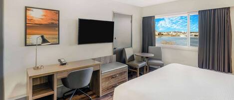A hotel room with a bed, desk, chair, TV, and a view of the city through the window.