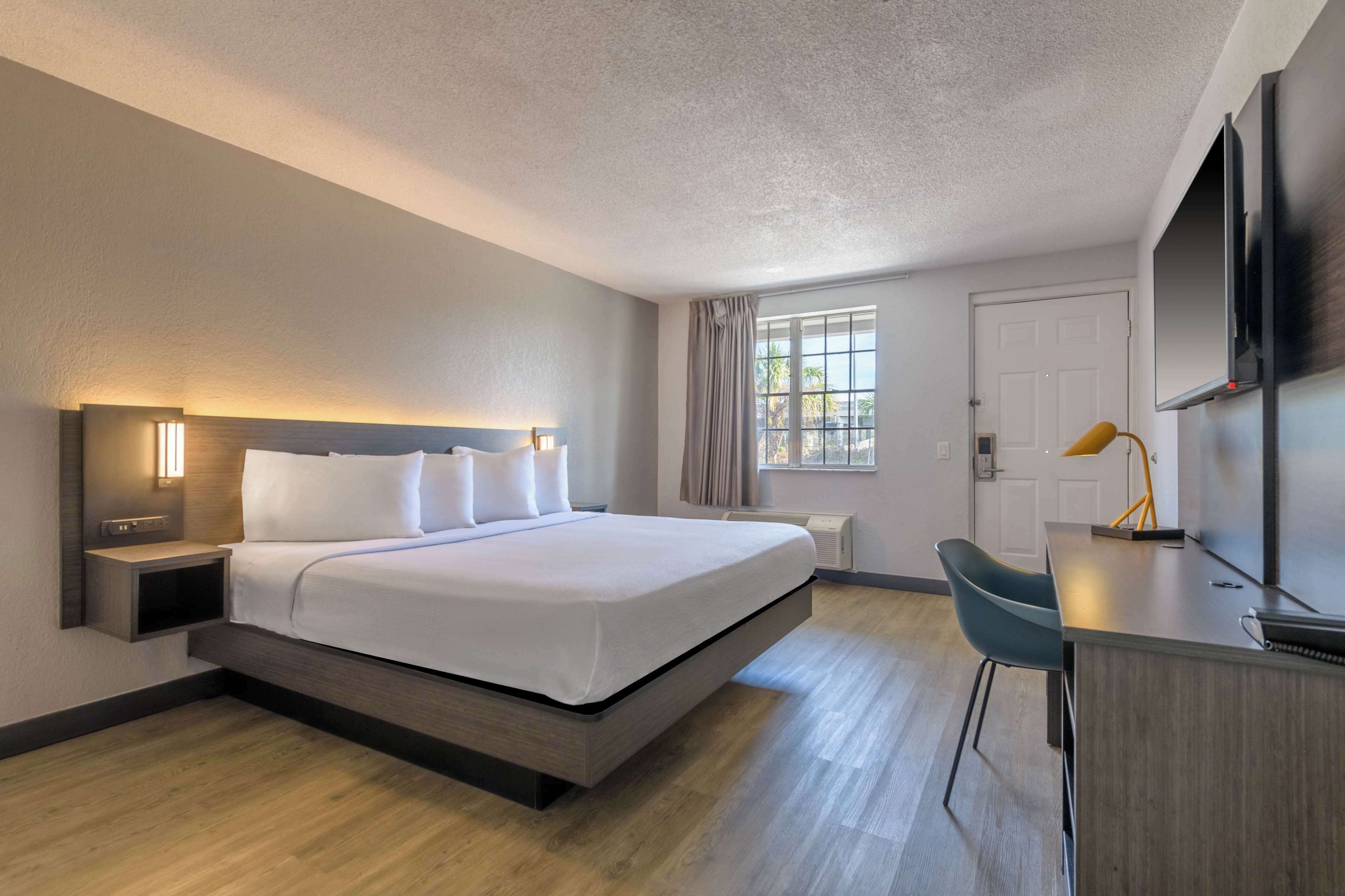 A modern hotel room with a bed, bedside table, lamp, desk, chair, and a TV mounted on the wall.