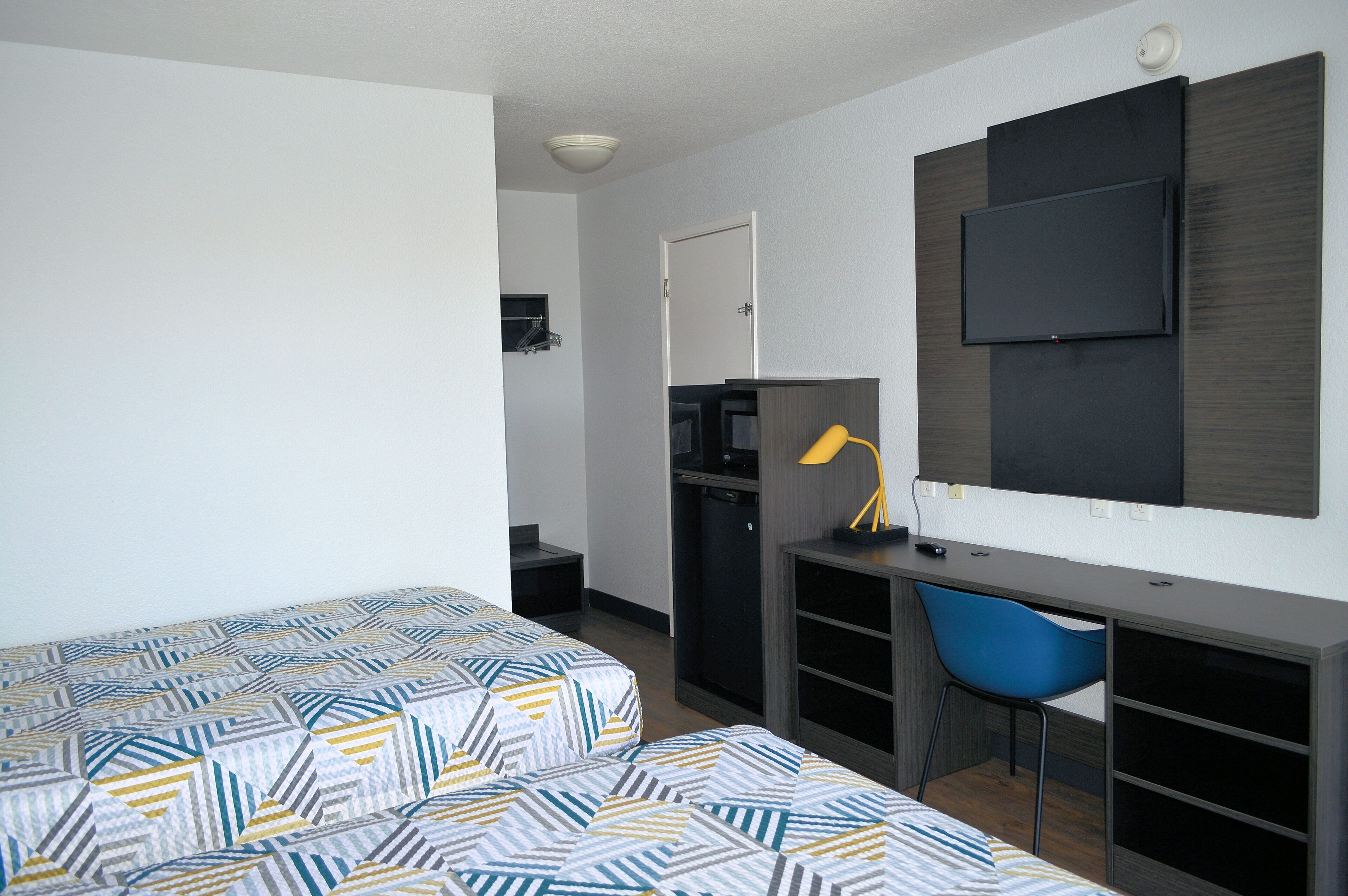 A compact hotel room with a bed, a desk, a chair, a television, and a small kitchenette.