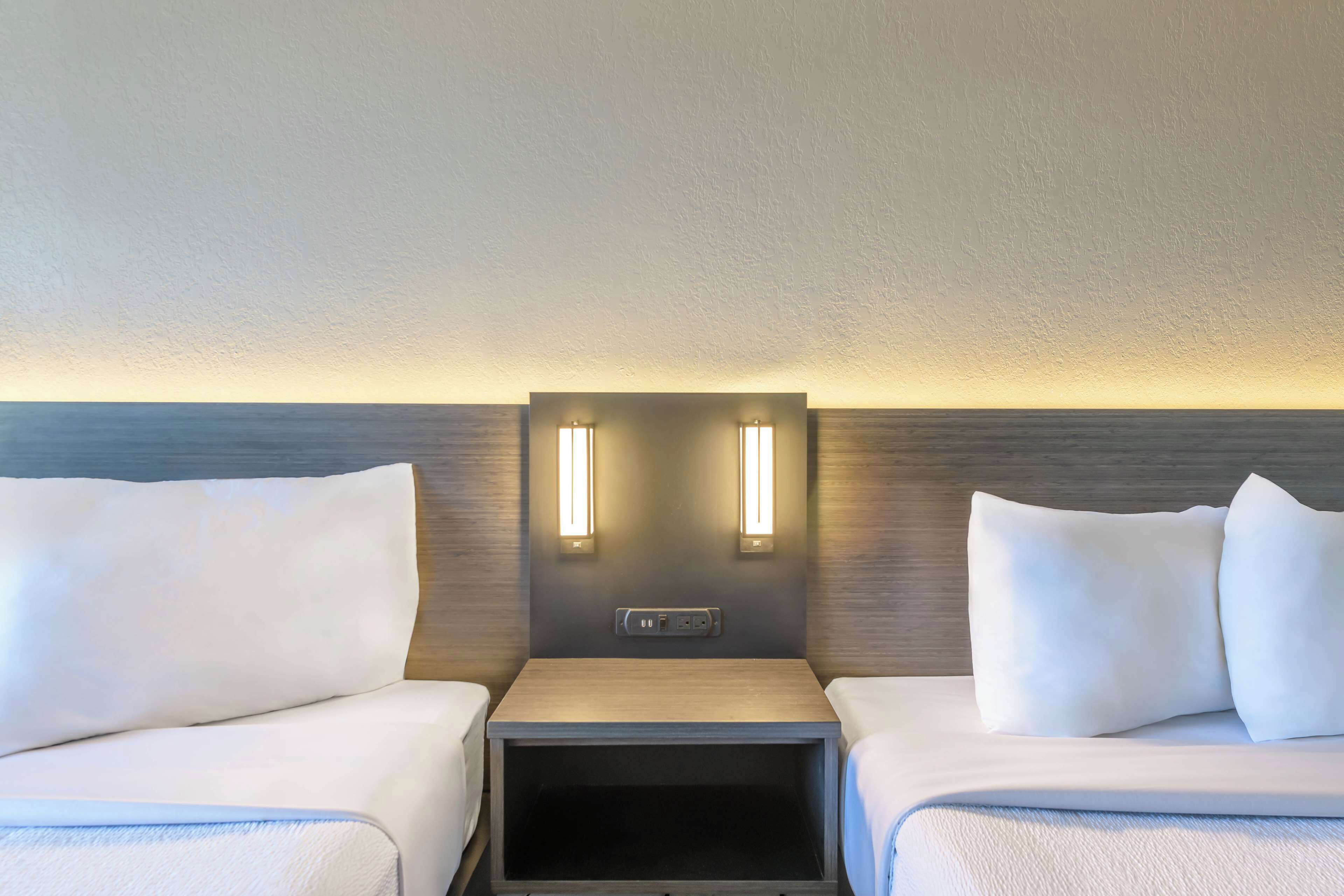 A hotel room with two beds, a nightstand, and a wall-mounted control panel.
