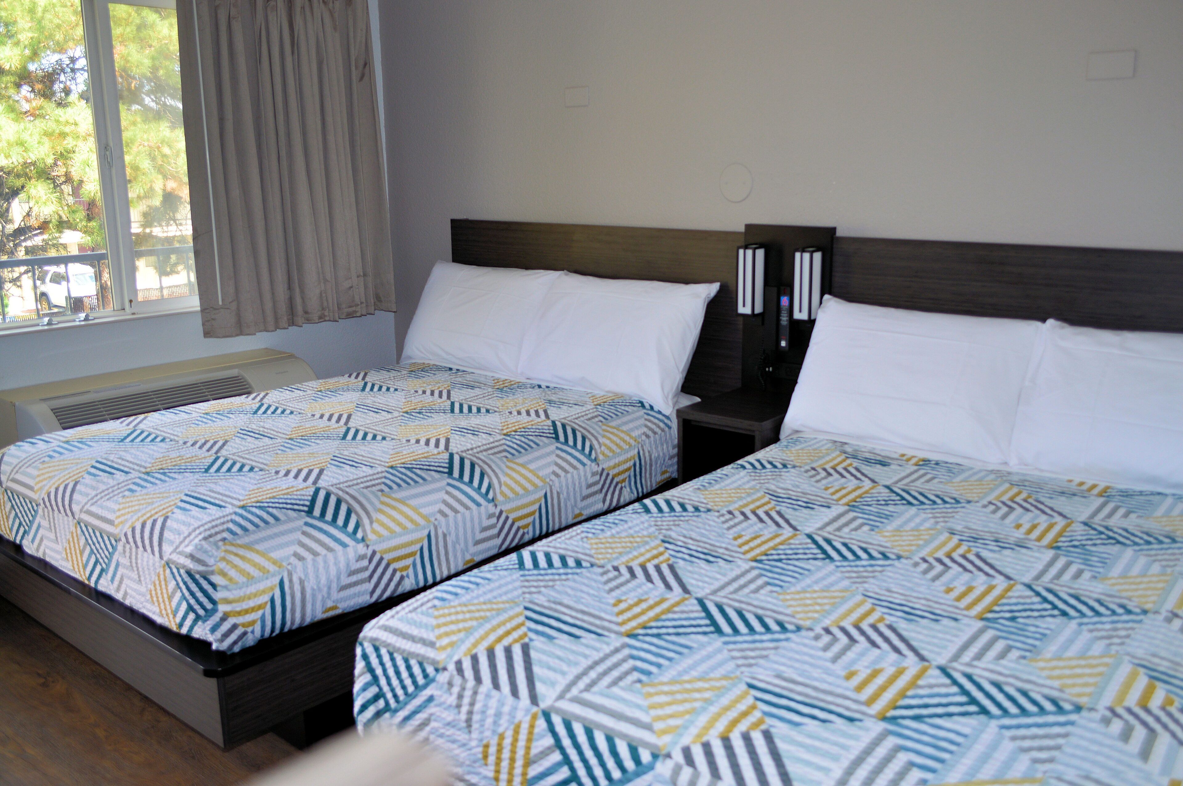 A hotel room with two beds, each with a patterned bedspread, a bedside table with a lamp, and a window with curtains.