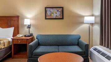 A hotel room with a bed, a sofa, a small table, a wall-mounted lamp, a heater, and a framed artwork on the wall.