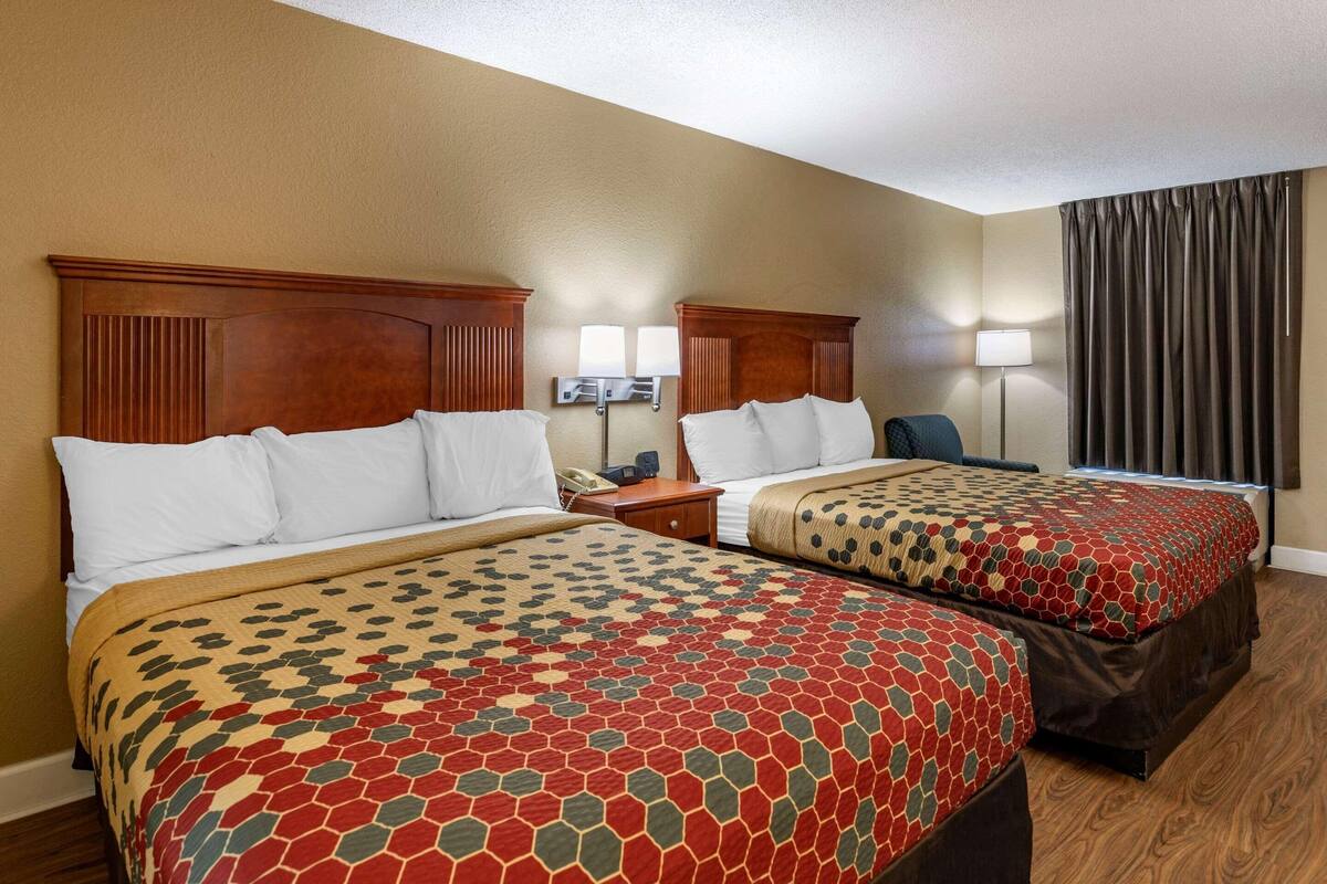 Ein Hotelzimmer mit zwei Betten, Holz-Kopfteilen, einem Schreibtisch, einem Stuhl, einer Lampe und einem Fenster mit Vorhängen.