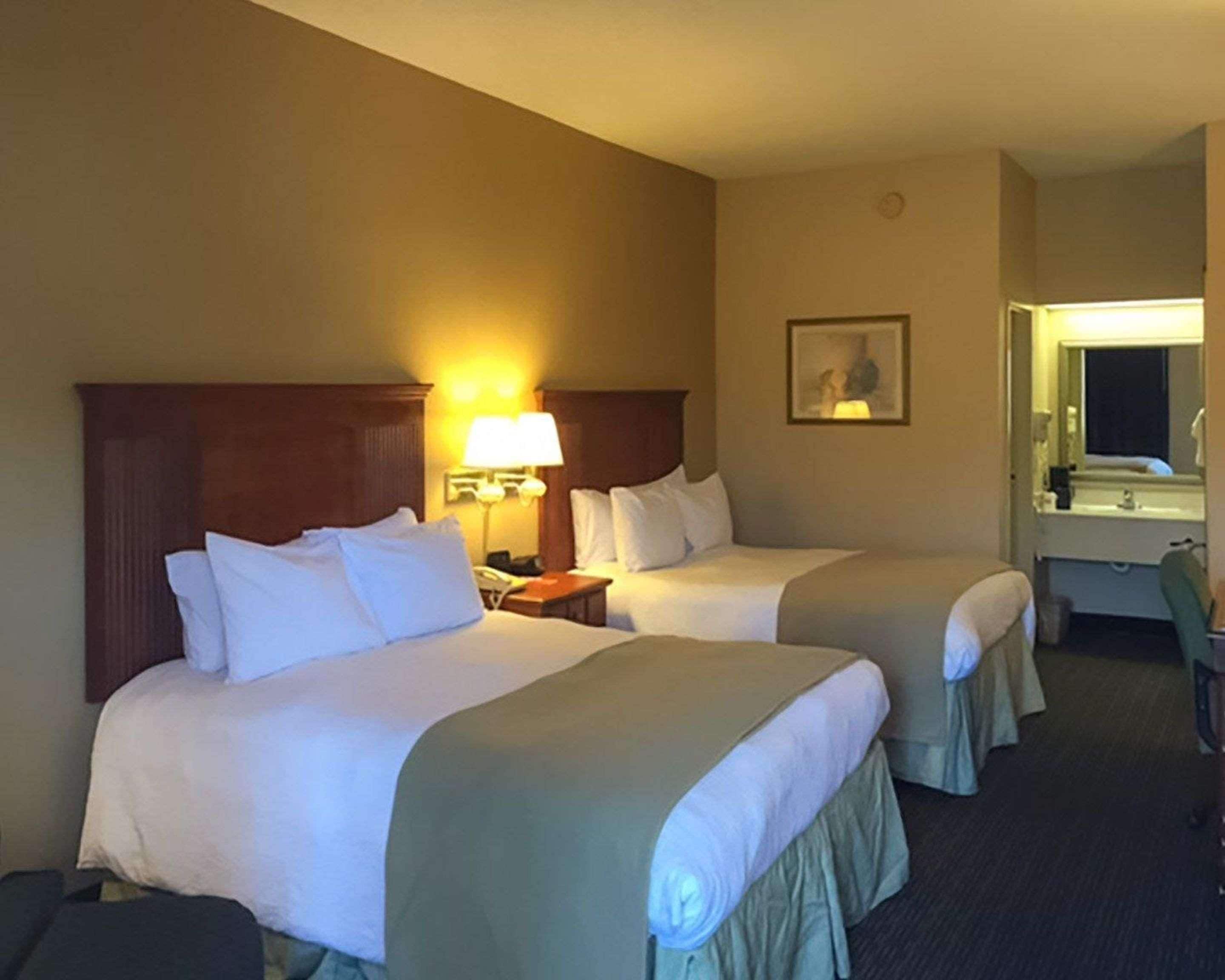 A hotel room with two beds, a nightstand with a lamp, a framed picture on the wall, and a bathroom visible through an open door.
