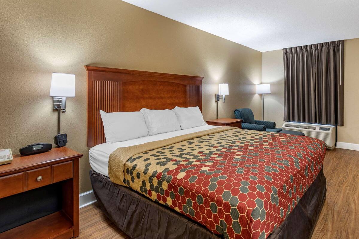 Ein Hotelzimmer mit Bett, zwei Nachttischen, einem Sessel, einem Schreibtisch, einer Lampe und einem Vorhangfenster.