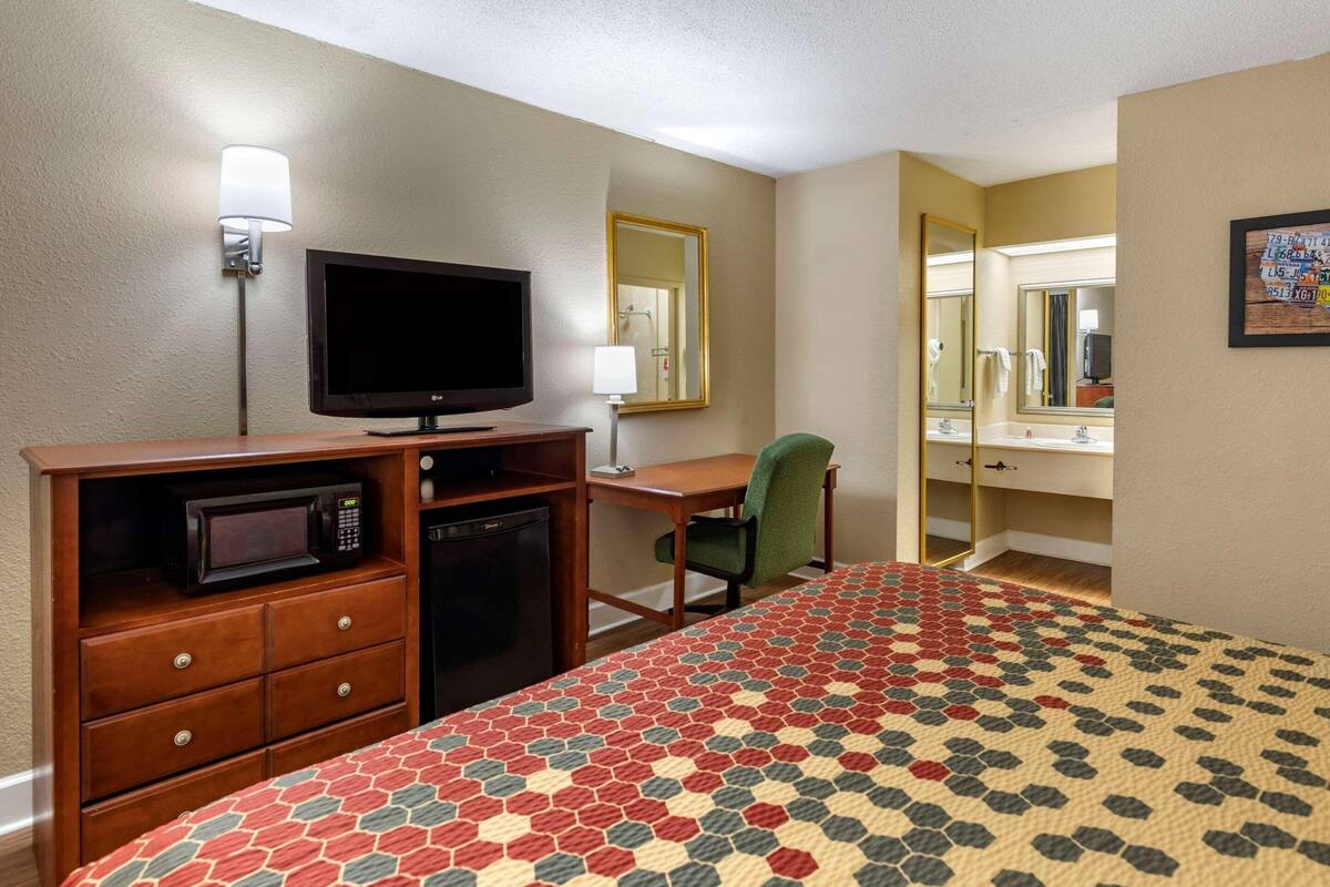 Ein Hotelzimmer mit Bett, Schreibtisch, Fernseher, Mikrowelle, Stuhl und Spiegel.