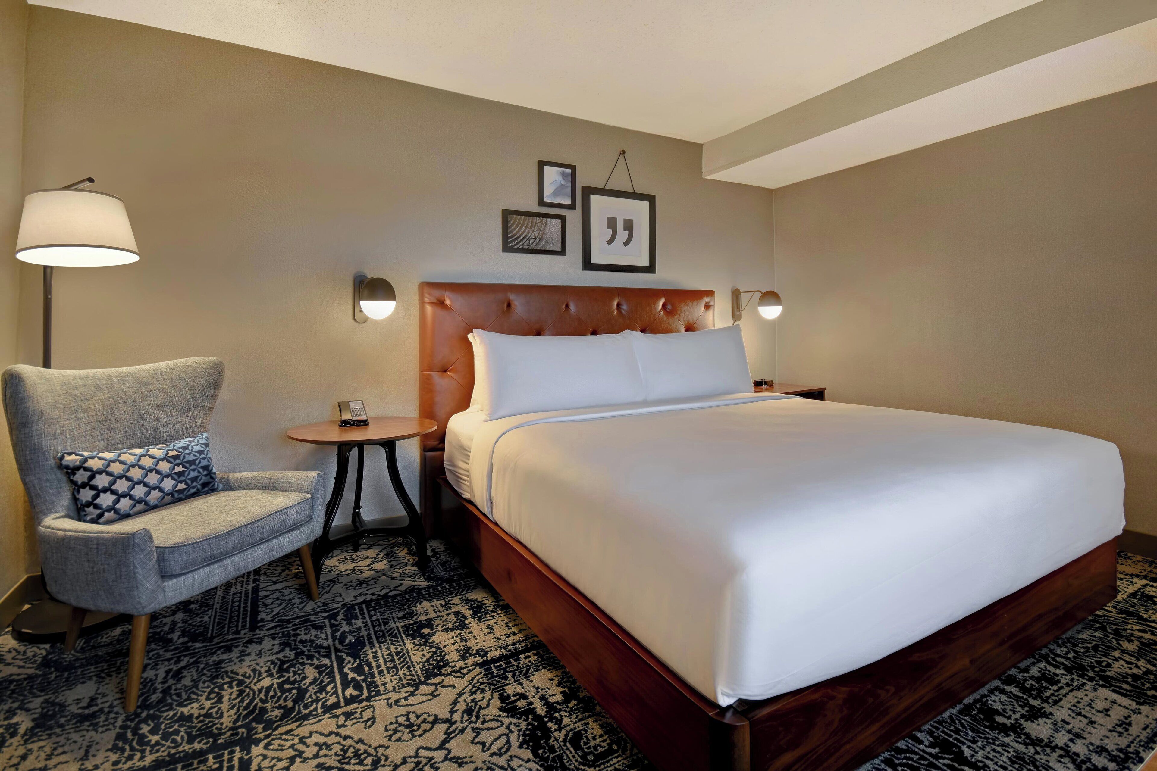 Una habitación de hotel con una cama grande, una silla, una mesita y lámparas fijadas a la pared.
