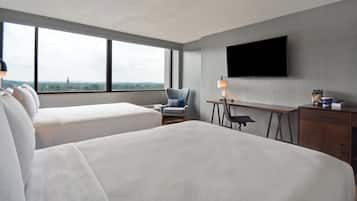 A hotel room with two beds, a desk, a chair, a TV, and a view of a cityscape.