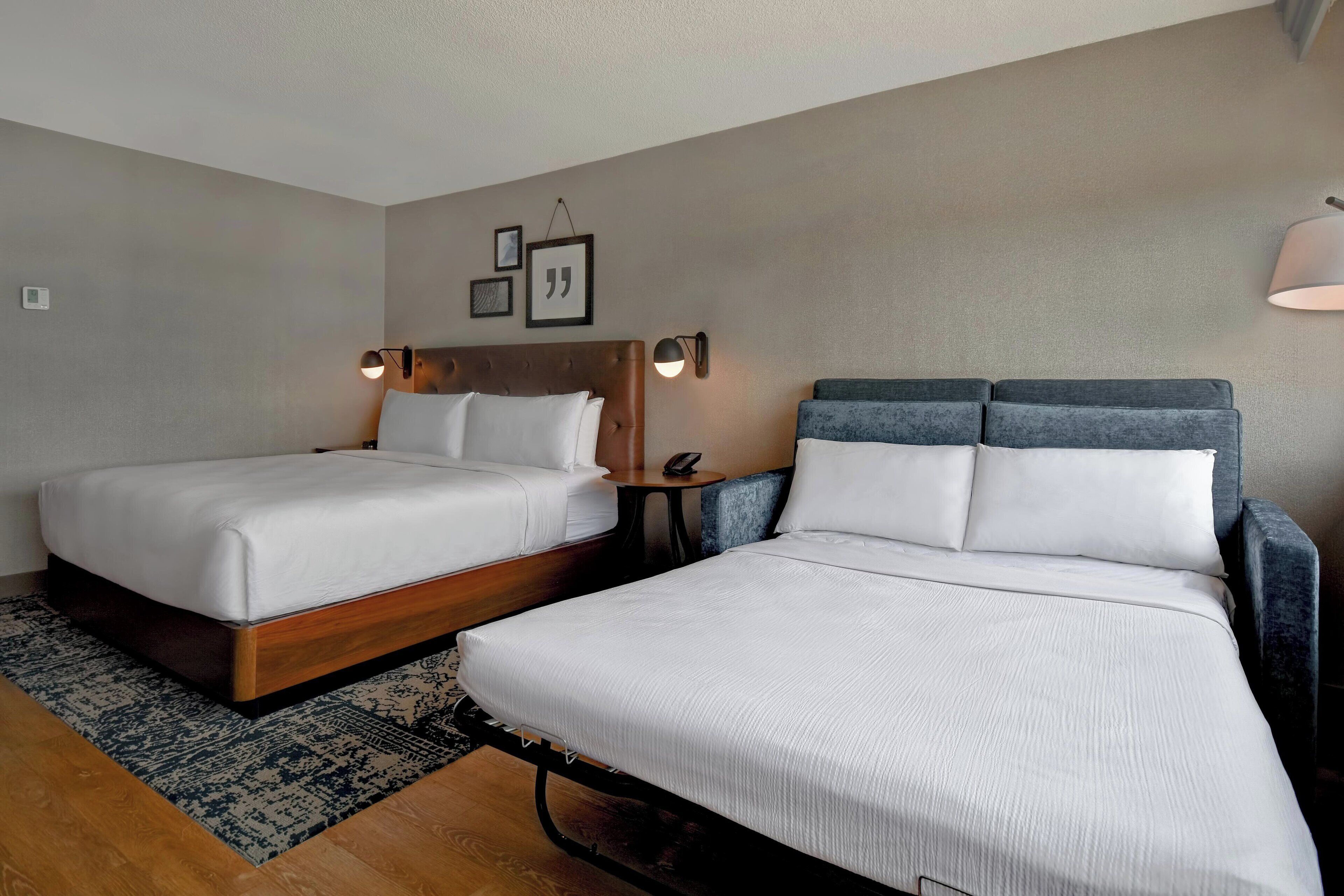 A hotel room with two beds, a small table, and a wall-mounted lamp.