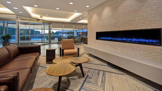A modern lobby with a leather sofa, two round wooden tables, a flat-screen TV mounted on a textured wall, and a glass partition with a view of the outdoors. - Four Points by Sheraton Boston Newton (Newton)
