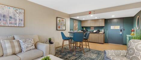 Una sala de estar moderna con un sofá, un sillón estampado, un comedor con sillas, una cocina con armarios de madera y un cuadro abstracto grande en la pared.