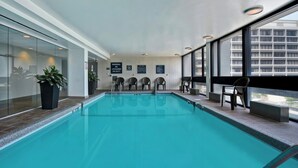 An indoor swimming pool with clear glass walls, a tiled floor, and several chairs and tables along the side. - Four Points by Sheraton Boston Newton (Newton)