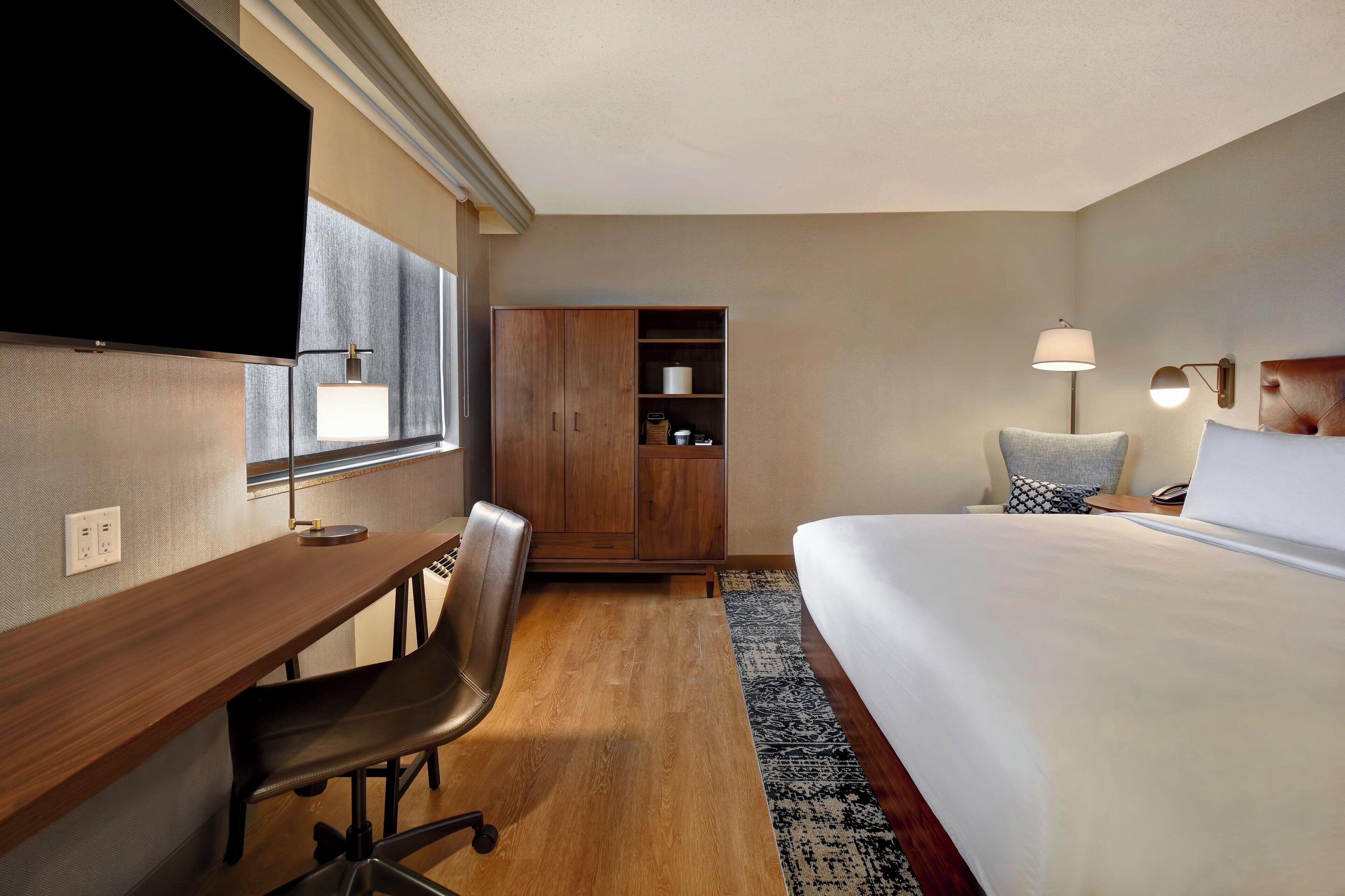 A hotel room with a bed, desk, chair, wardrobe, TV, lamp, and a nightstand.