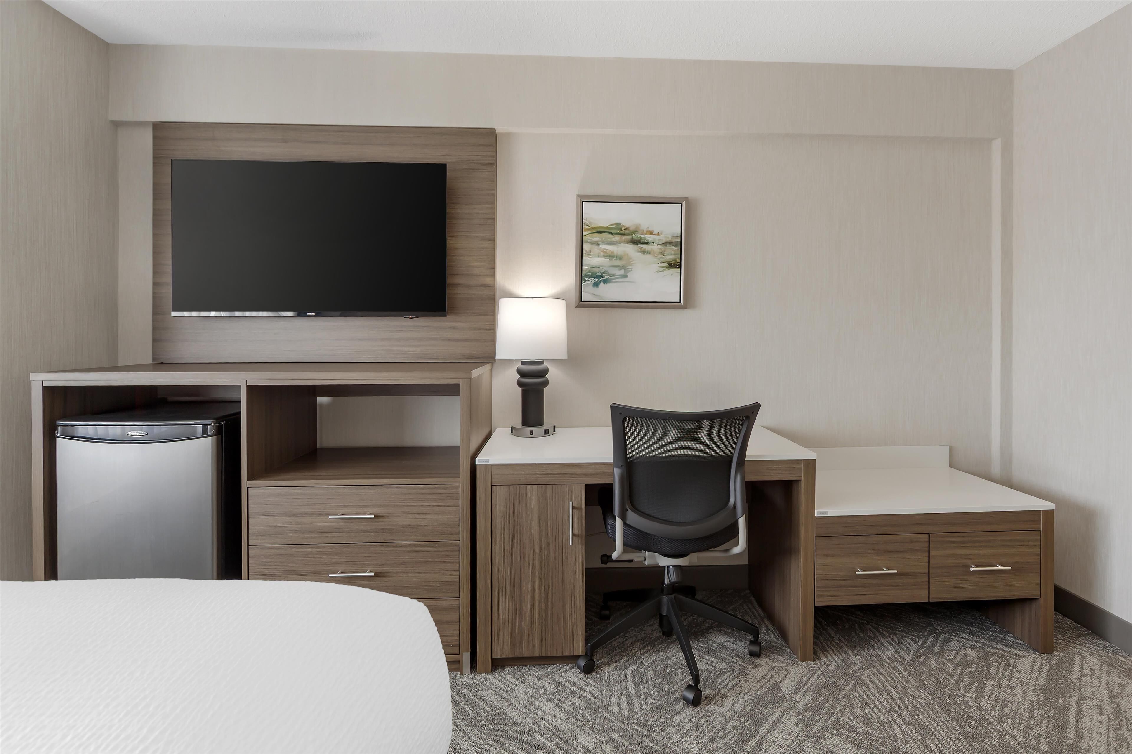 Una habitación de hotel con cama, escritorio con silla, televisor, refrigerador y lámpara.