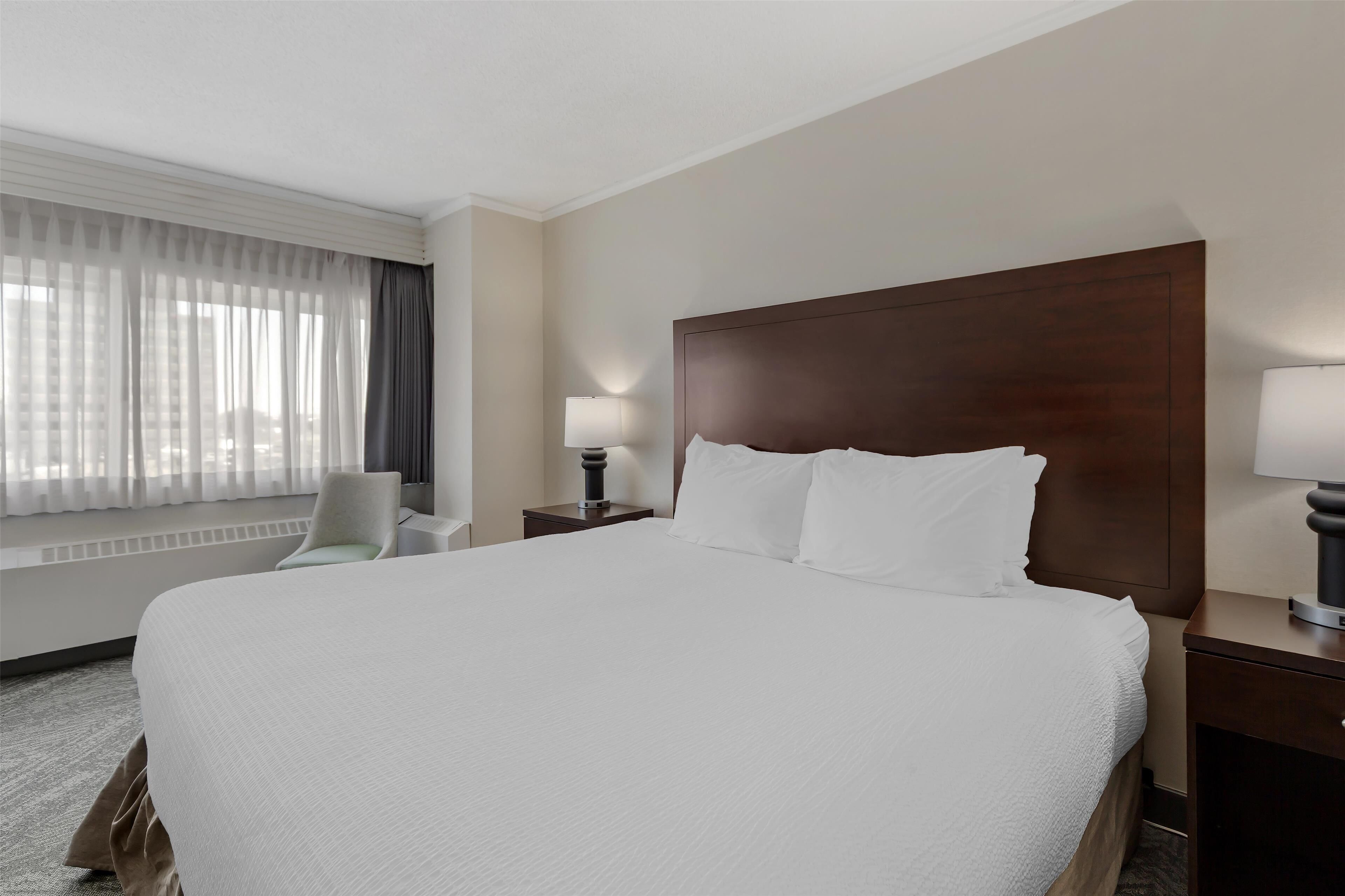 A hotel room with a large bed, a nightstand with a lamp, a chair, and a window with curtains.