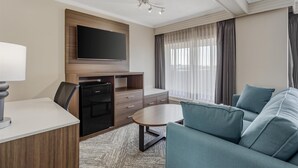 A modern hotel room with a flat-screen TV mounted on a wooden panel, a dining area with a table and chairs, a sofa, and a large window with curtains. - Best Western Cedar Park Inn (Edmonton)