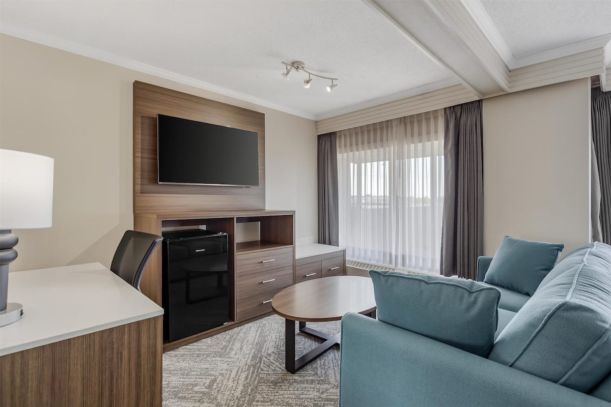 A modern hotel room with a flat-screen TV mounted on a wooden panel, a dining area with a table and chairs, a sofa, and a large window with curtains.