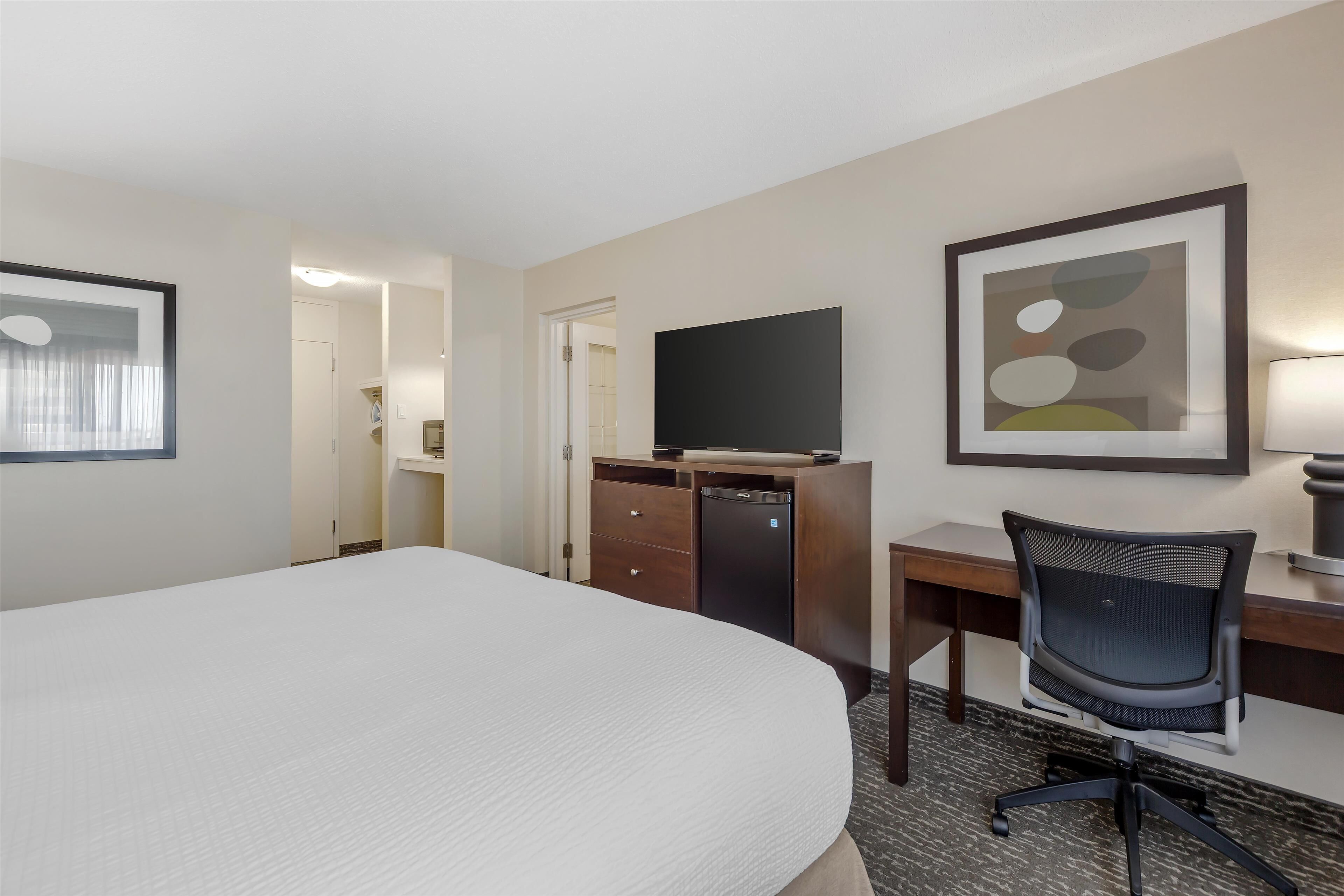 A hotel room with a large bed, a desk with a chair, a flat-screen TV, a dresser, and a framed abstract art piece on the wall.
