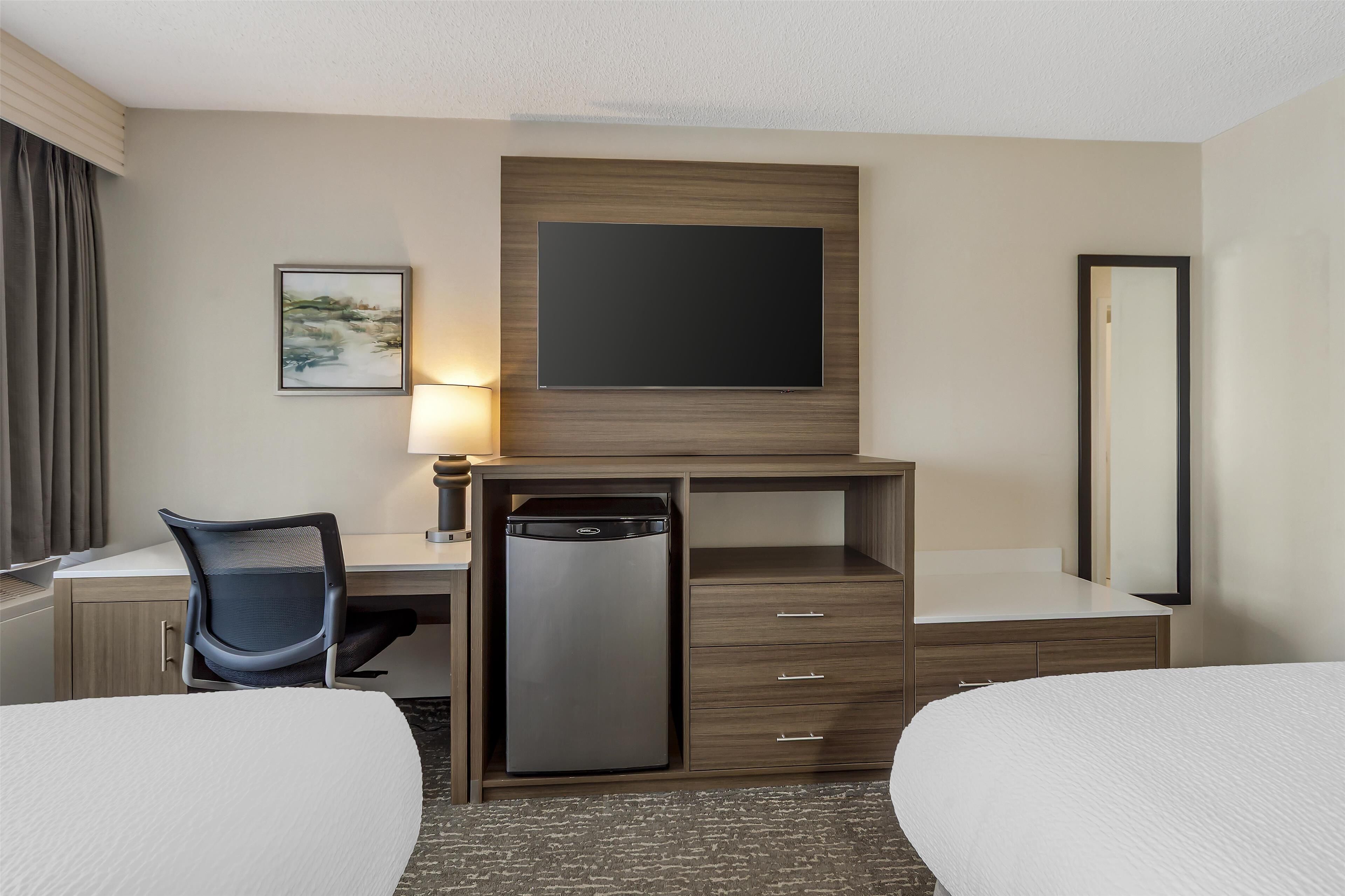 A hotel room with a bed, a desk with a chair, a small refrigerator, a flat-screen TV mounted on the wall, a bedside table, and a mirror.