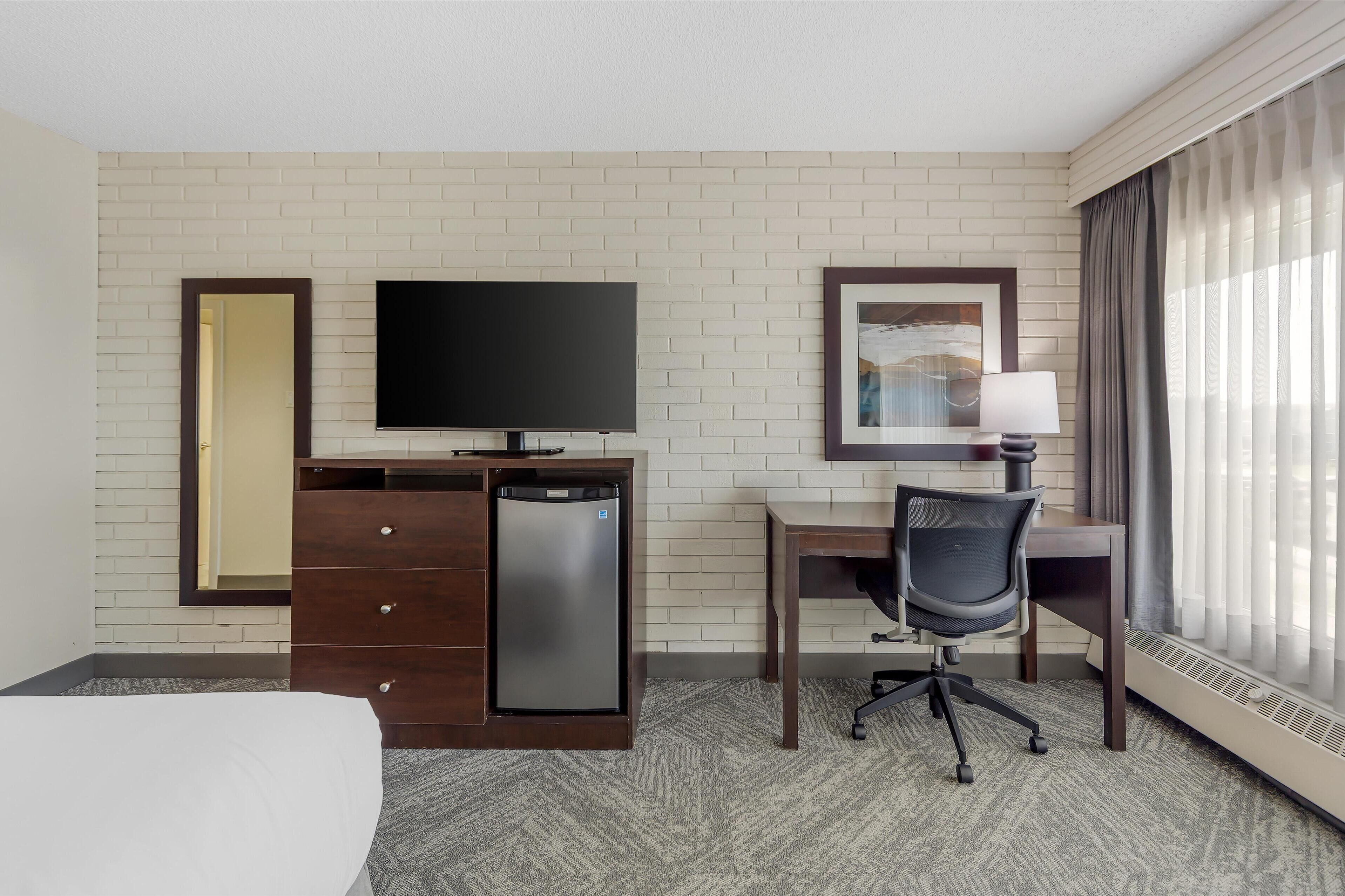 A hotel room with a bed, a desk with a chair, a television, a mini-fridge, a dresser, a lamp, and a window with curtains.