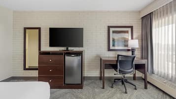 A hotel room with a bed, a desk with a chair, a television, a mini-fridge, a dresser, a lamp, and a window with curtains.