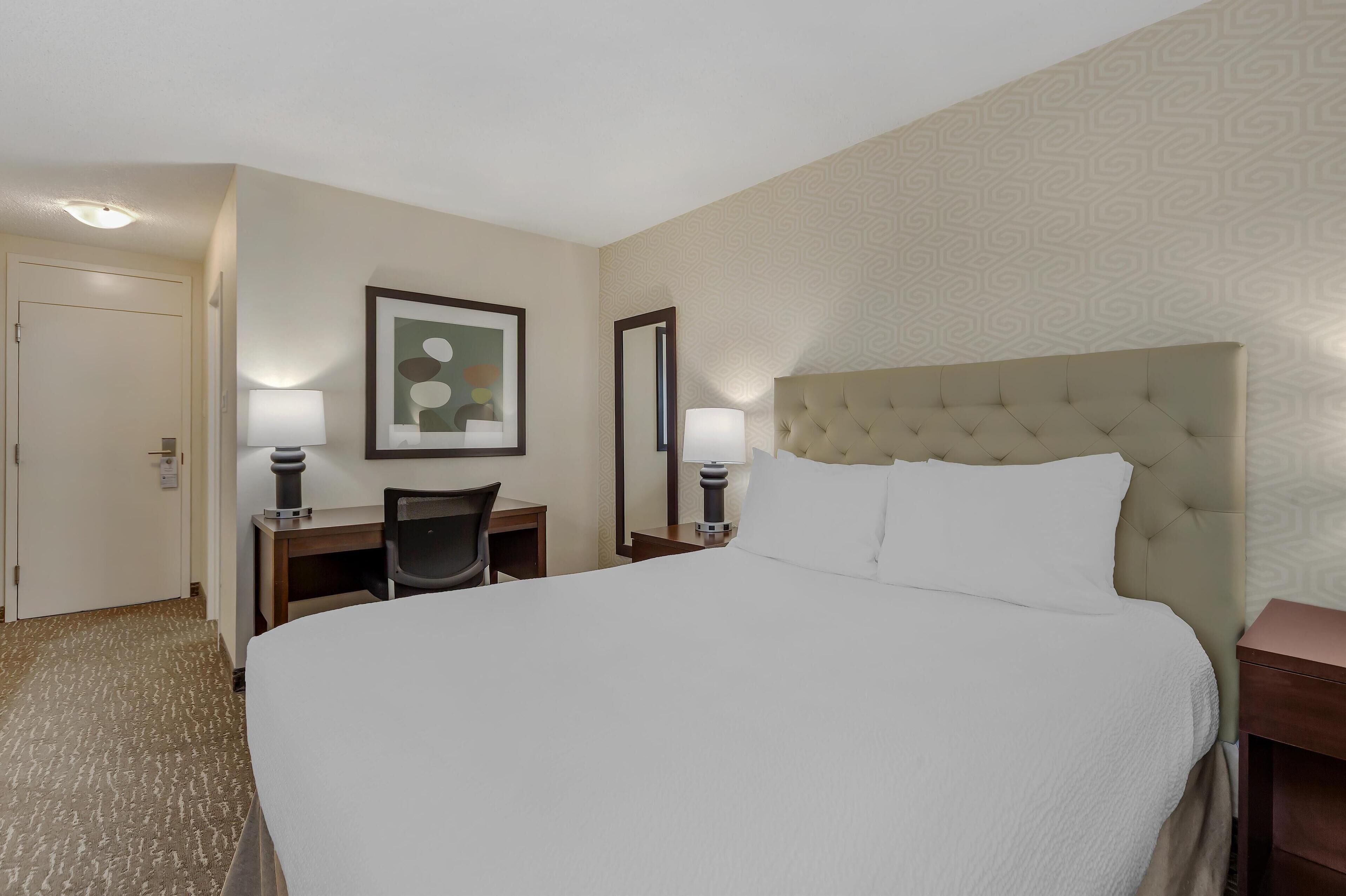 A hotel room with a large bed, a desk with a chair, two bedside lamps, a framed picture on the wall, and a door.