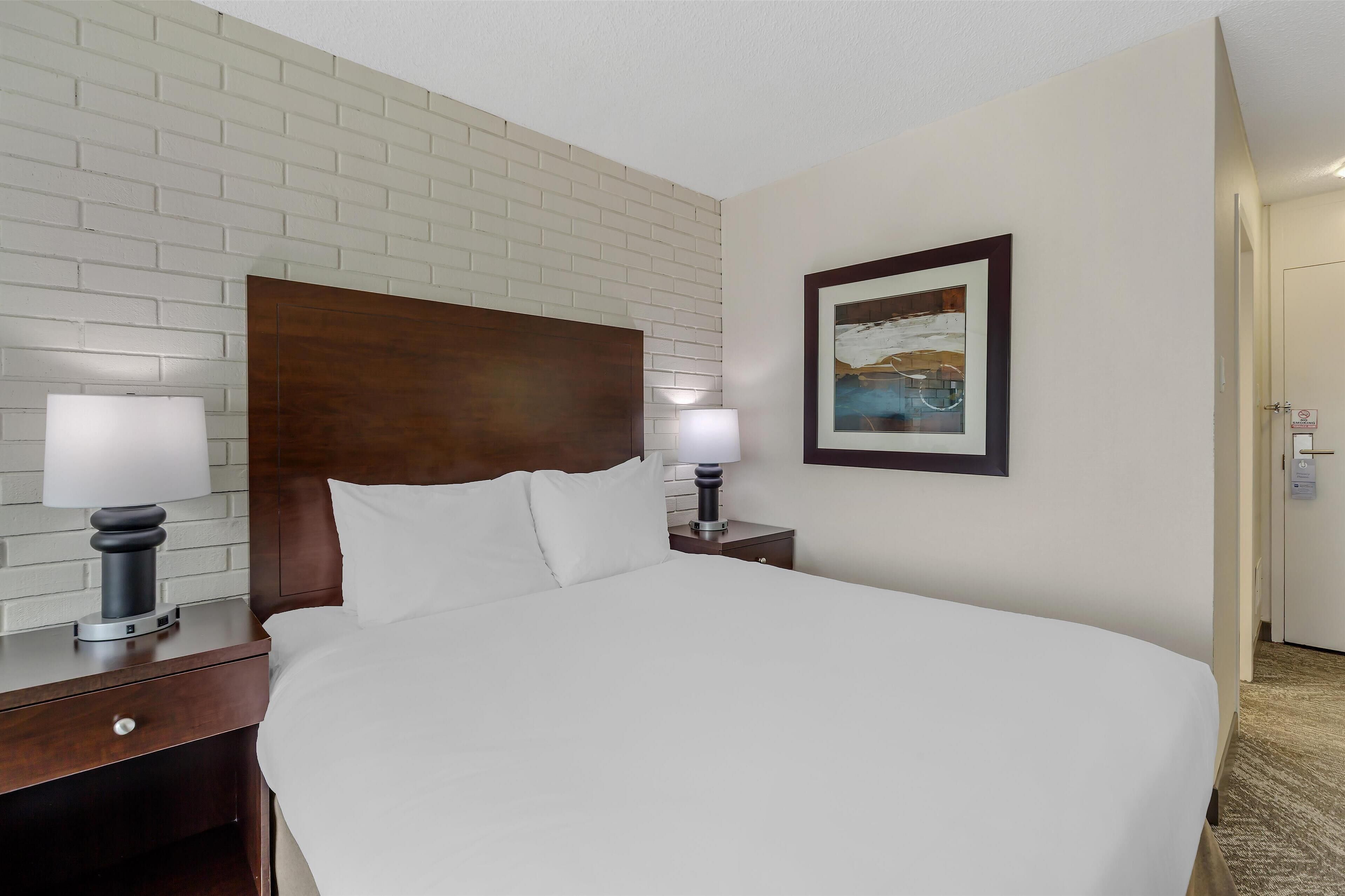 A hotel room with a large bed, two bedside tables with lamps, a wooden headboard, a framed picture on the wall, and a door to the right.