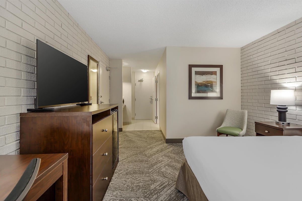 A hotel room with a bed, a desk with a flat-screen TV, a dresser, a chair, and a nightstand with a lamp.