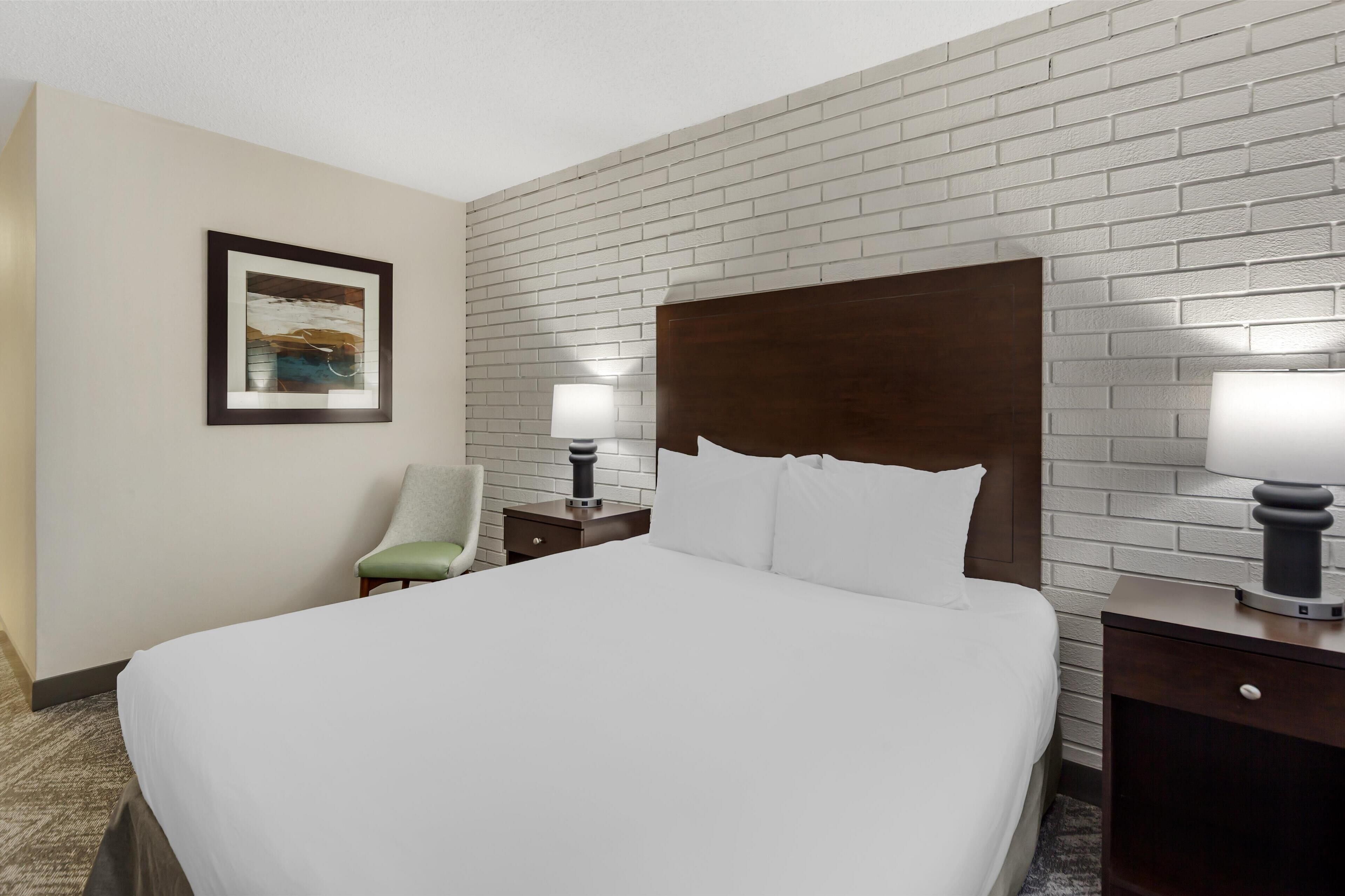 A hotel room with a large bed, a nightstand with a lamp, a chair, and a framed picture on the wall.
