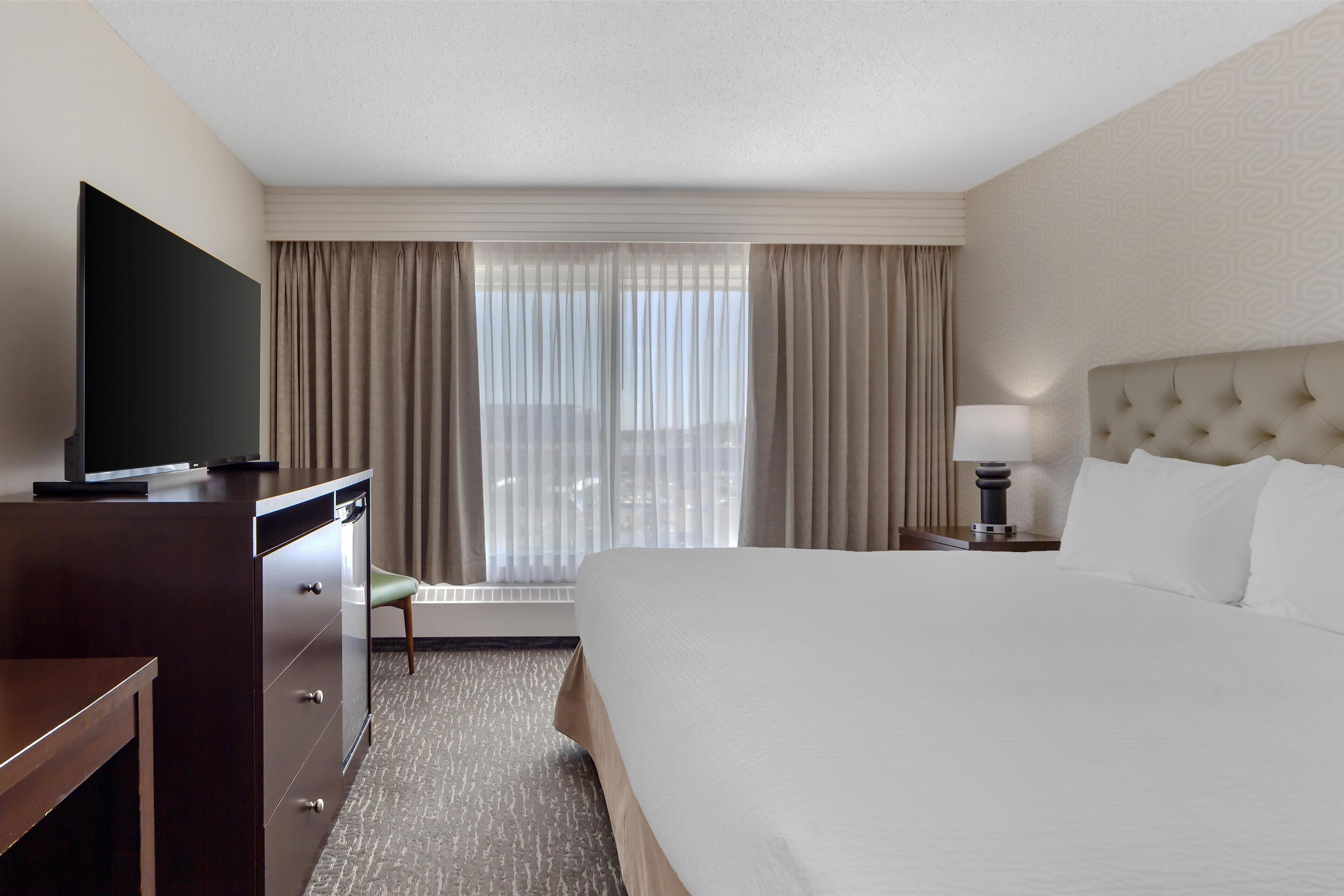 A hotel room with a large bed, a television on a dresser, a desk, and a window with curtains.