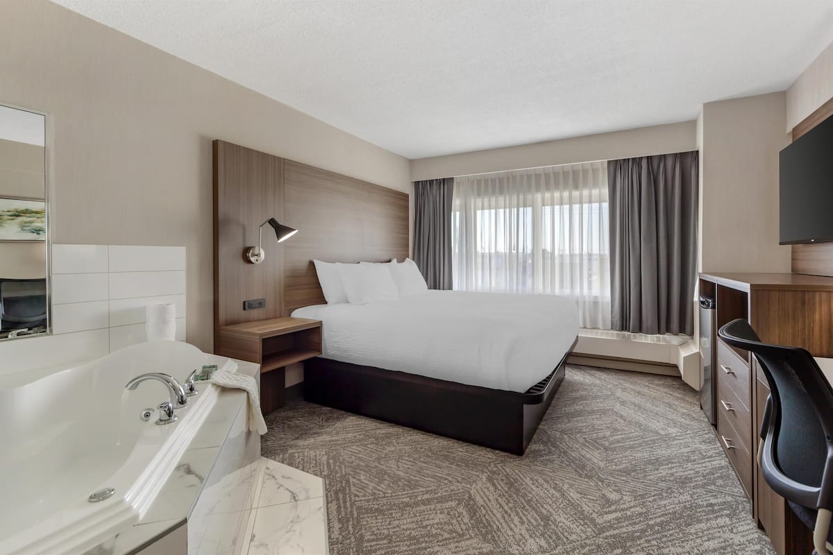 A modern hotel room with a large bed, a separate desk area, a bathtub, and a large window with curtains.