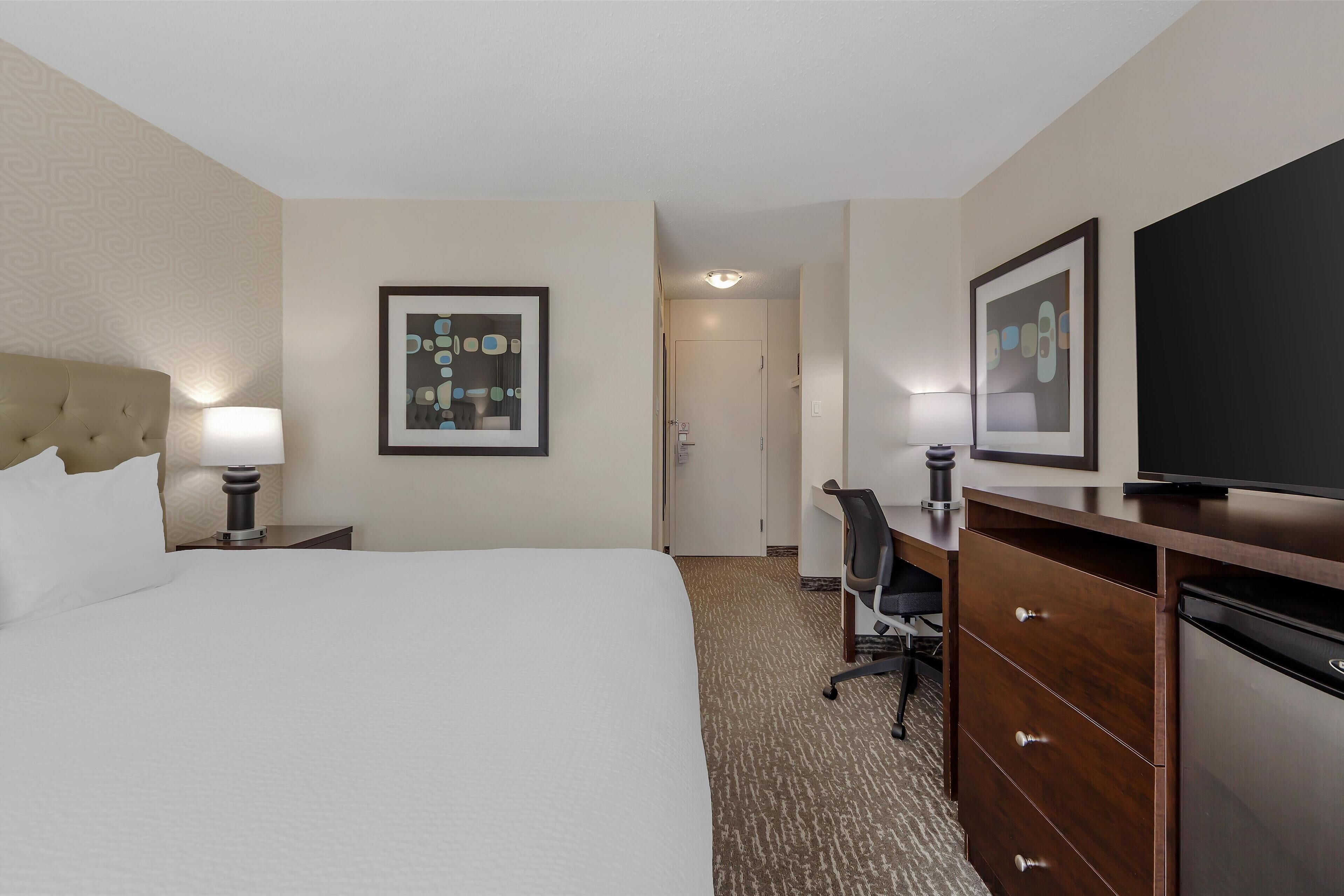 A hotel room with a large bed, a desk with a chair, a dresser, a television, and a framed picture on the wall.