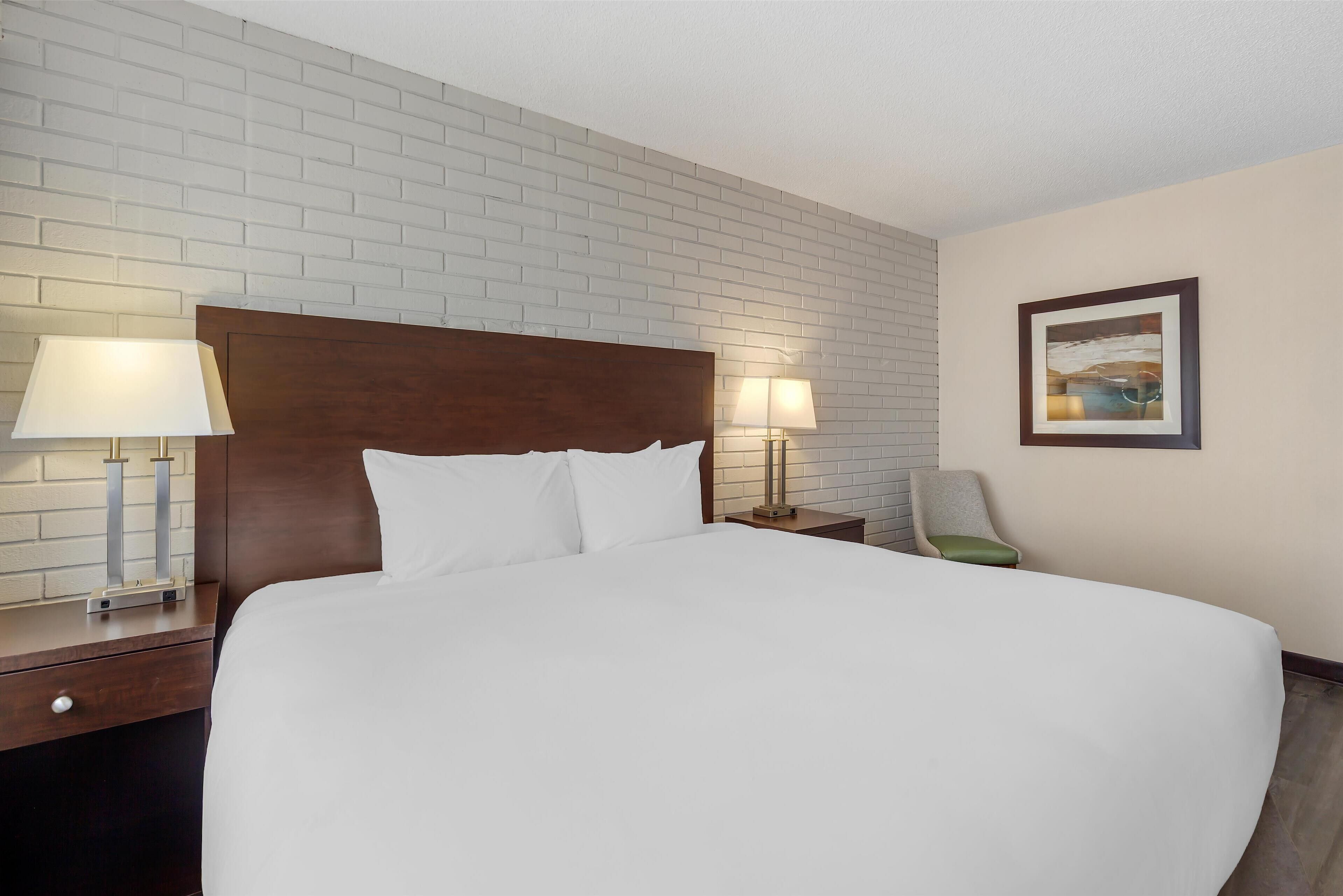 Habitación de hotel con una cama grande, dos lámparas de noche, un cabecero de madera, una mesita de noche, una silla y un cuadro en la pared.