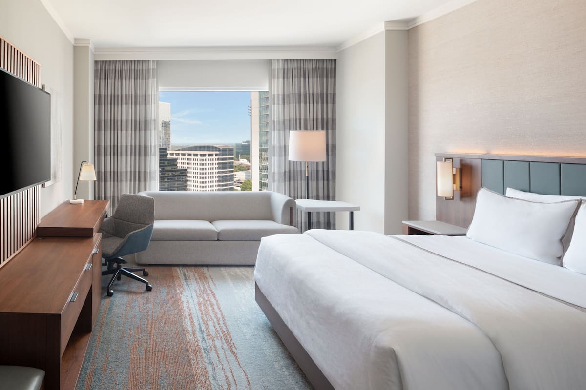 Habitación de hotel con una cama grande, un sofá, un escritorio con silla, un televisor y una ventana con cortinas.