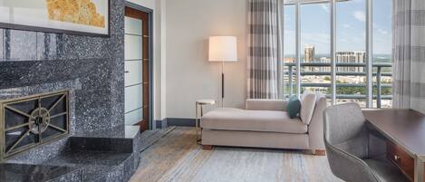 A modern hotel room with a curved marble fireplace, a sofa, a desk, and a large window offering a city view.