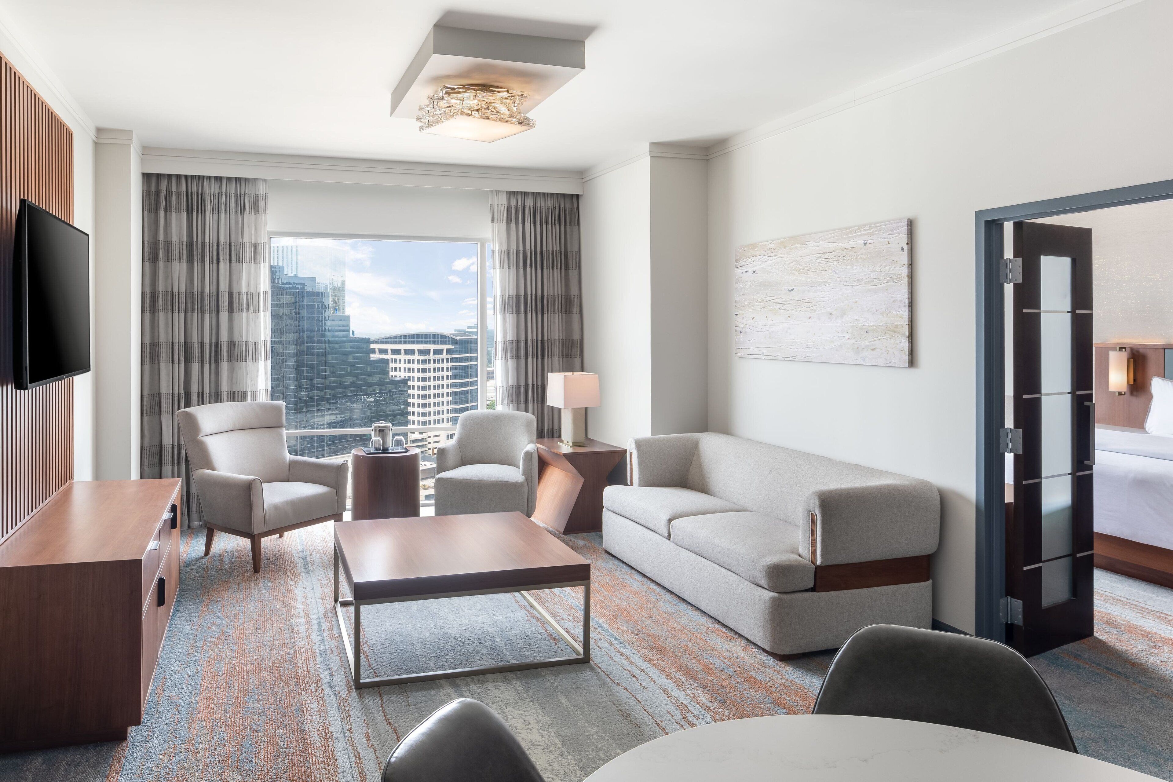 A modern hotel room with a large window, a flat-screen TV, a sofa, a coffee table, and a desk.