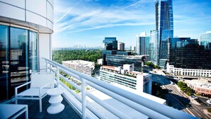 A balcony with a white chair and table, overlooking a cityscape with modern buildings and a clear sky.