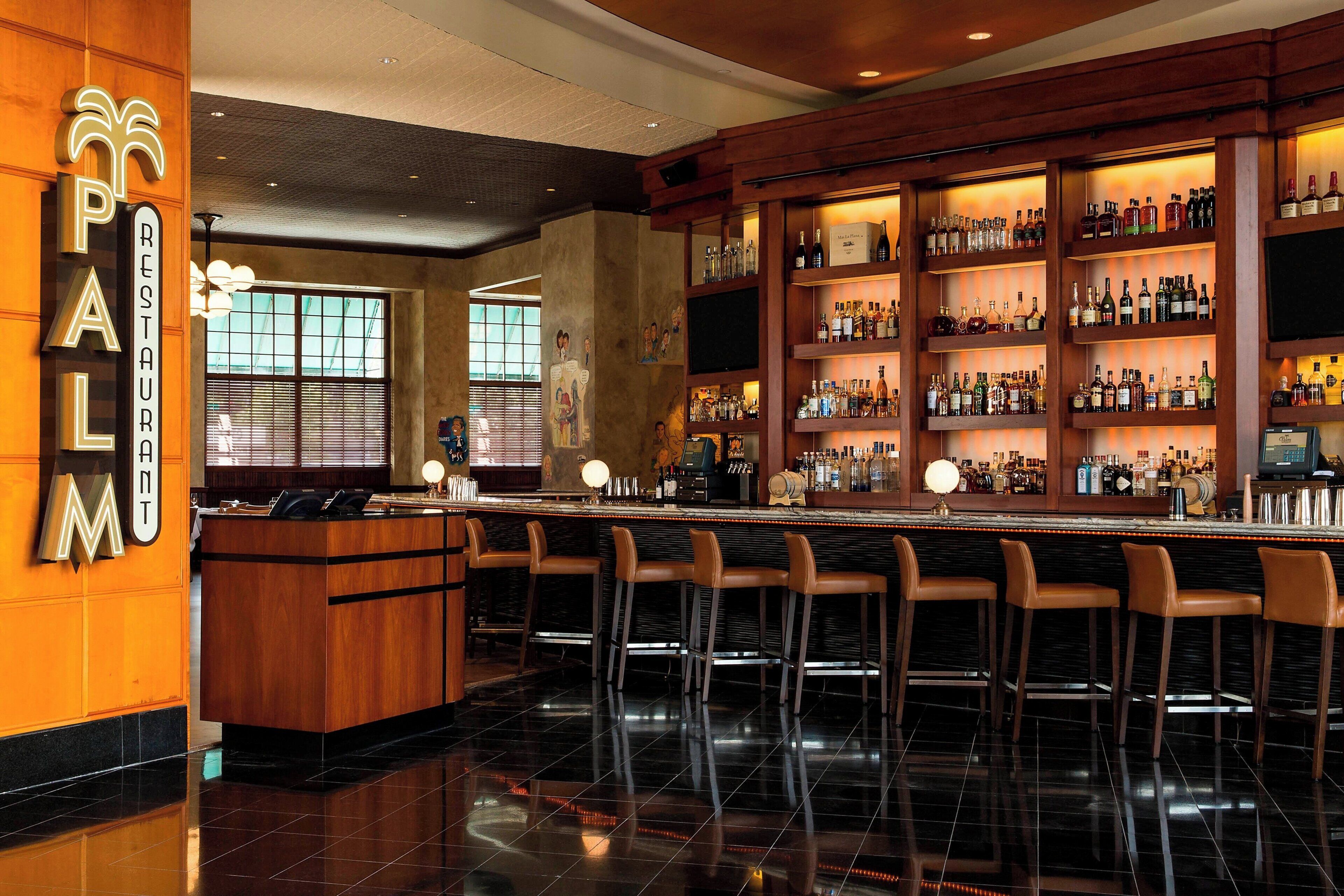 Una zona de bar con un mostrador de recepción de madera, varios taburetes y estanterías llenas de botellas.