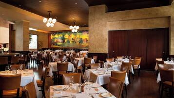 A spacious restaurant with multiple tables set for dining, featuring a large mural on the back wall and a modern chandelier.