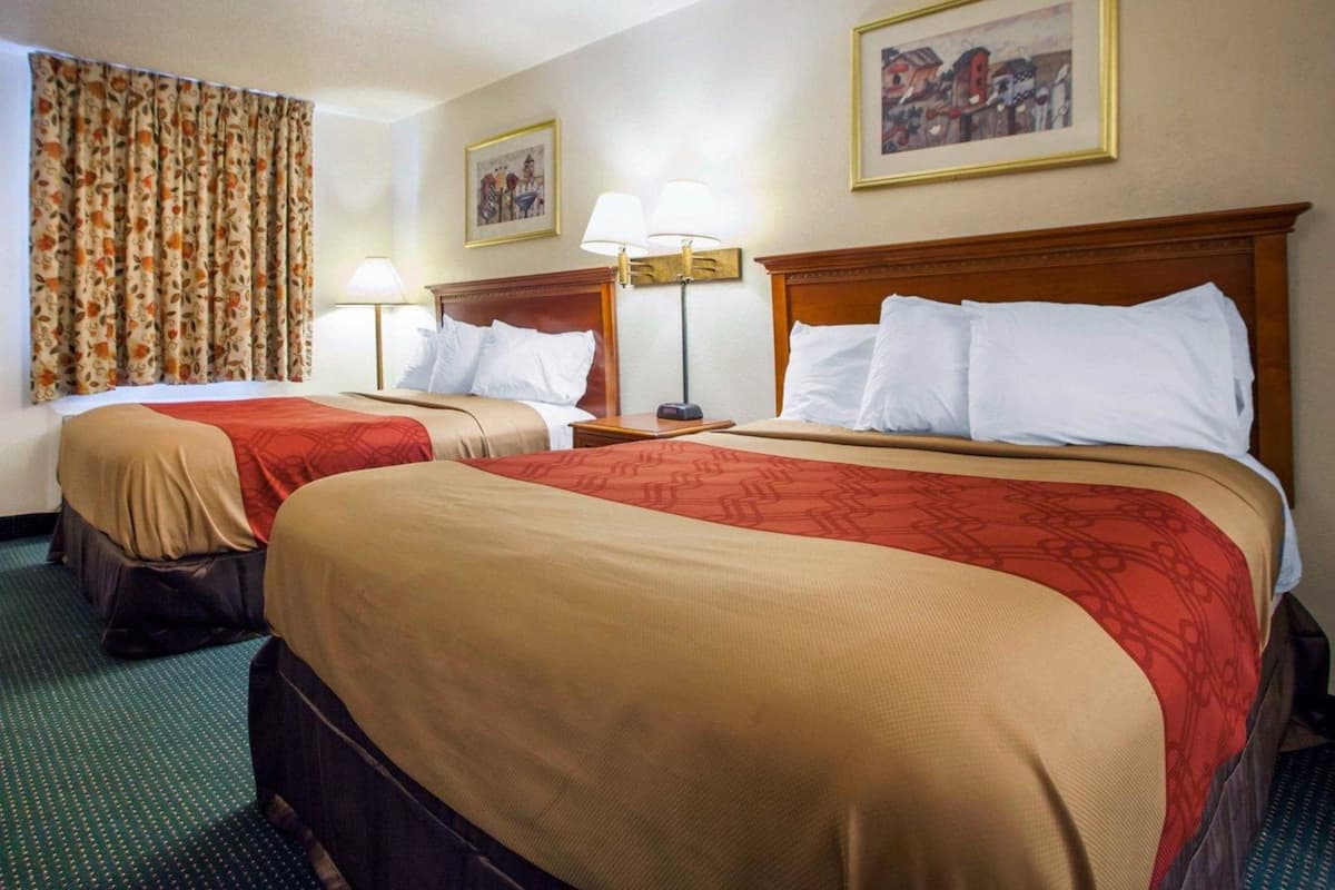 Quarto de hotel com duas camas, cada uma com cabeceira de madeira, colcha bege e vermelha, e tapete verde. Há duas luminárias de cabeceira e um quadro emoldurado na parede.