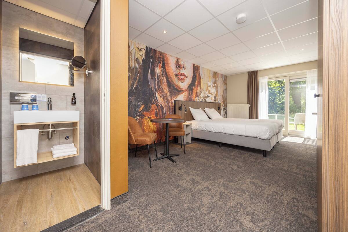 A hotel room with a bed, a round table, a sofa, a large painting on the wall, a bathroom with a mirror, and a window.
